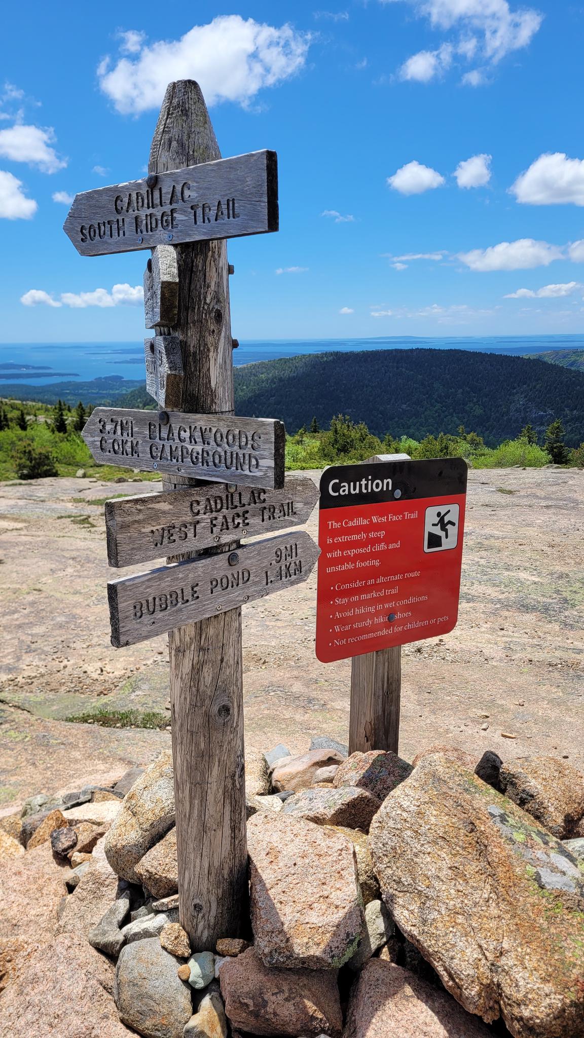 Wooden trail signpost on a rocky mountain top under a bright blue sky.