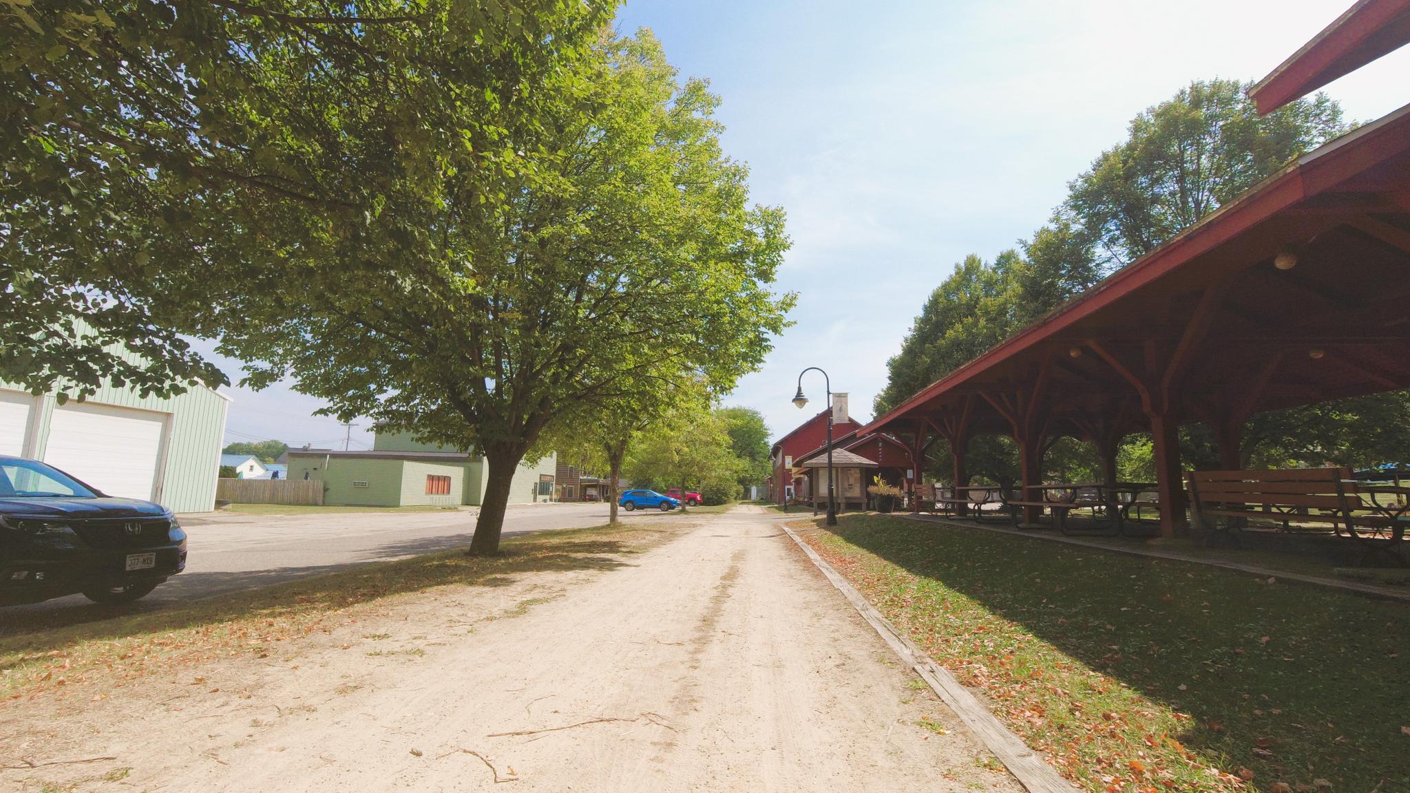 Dirt path leads past a red pavilion and green trees on a sunny day.