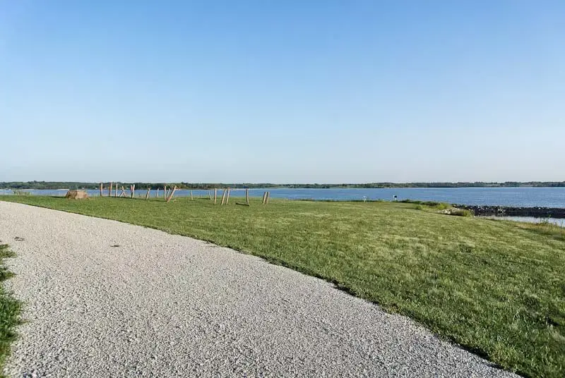 A sunny landscape with a gravel path, green lawn, and calm blue water.