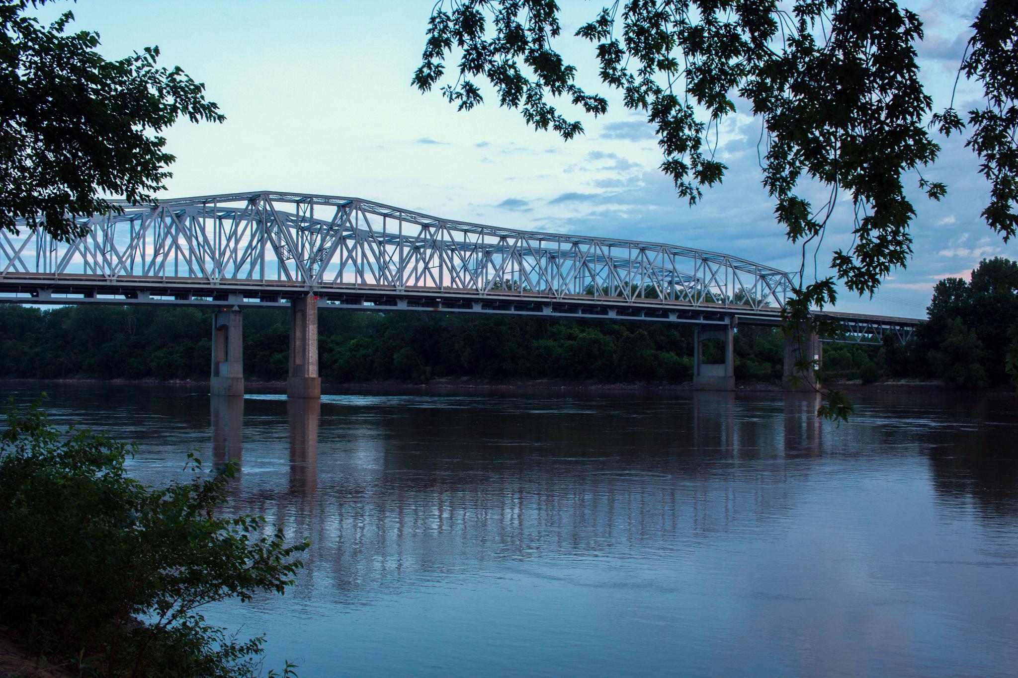 A large metal truss bridge spans a wide river at dusk.