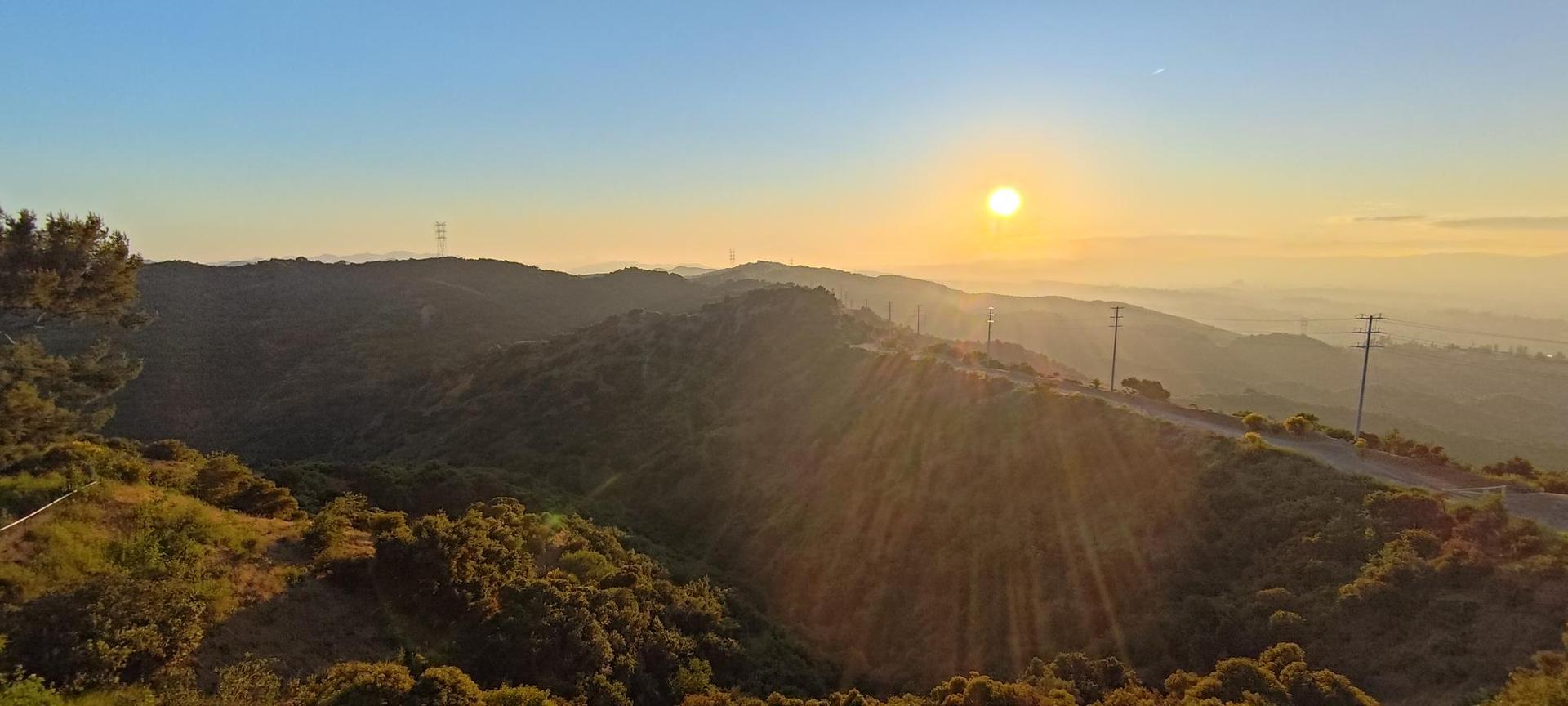 Golden sunset over rolling hills, sun rays, and power lines.