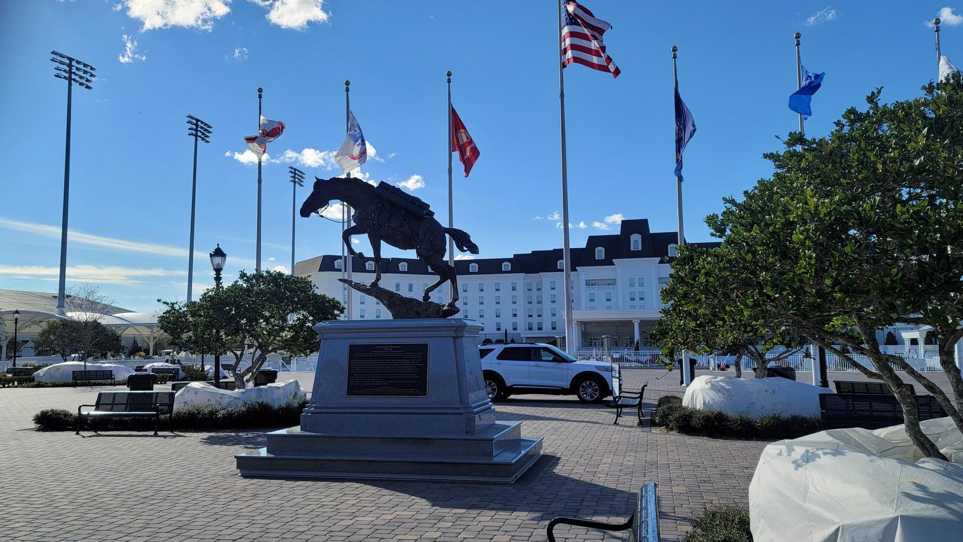 Horse statue in a plaza with flags, a large building, and blue sky.