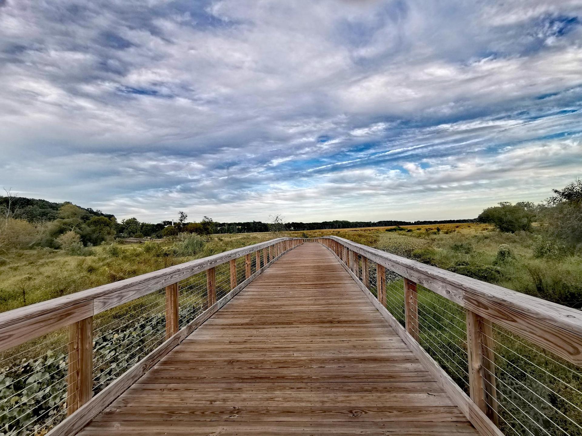 A long wooden boardwalk extends over a marsh under a dramatic cloudy sky.