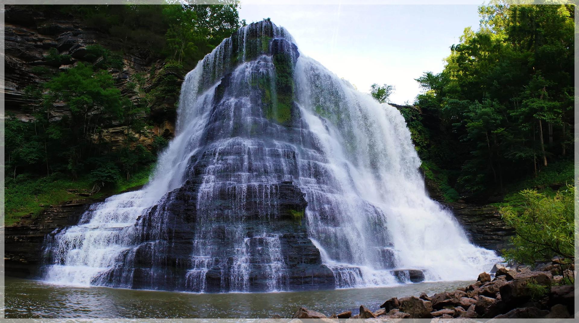 Massive multi-tiered waterfall plunging into a pool, framed by rocky cliffs and trees.