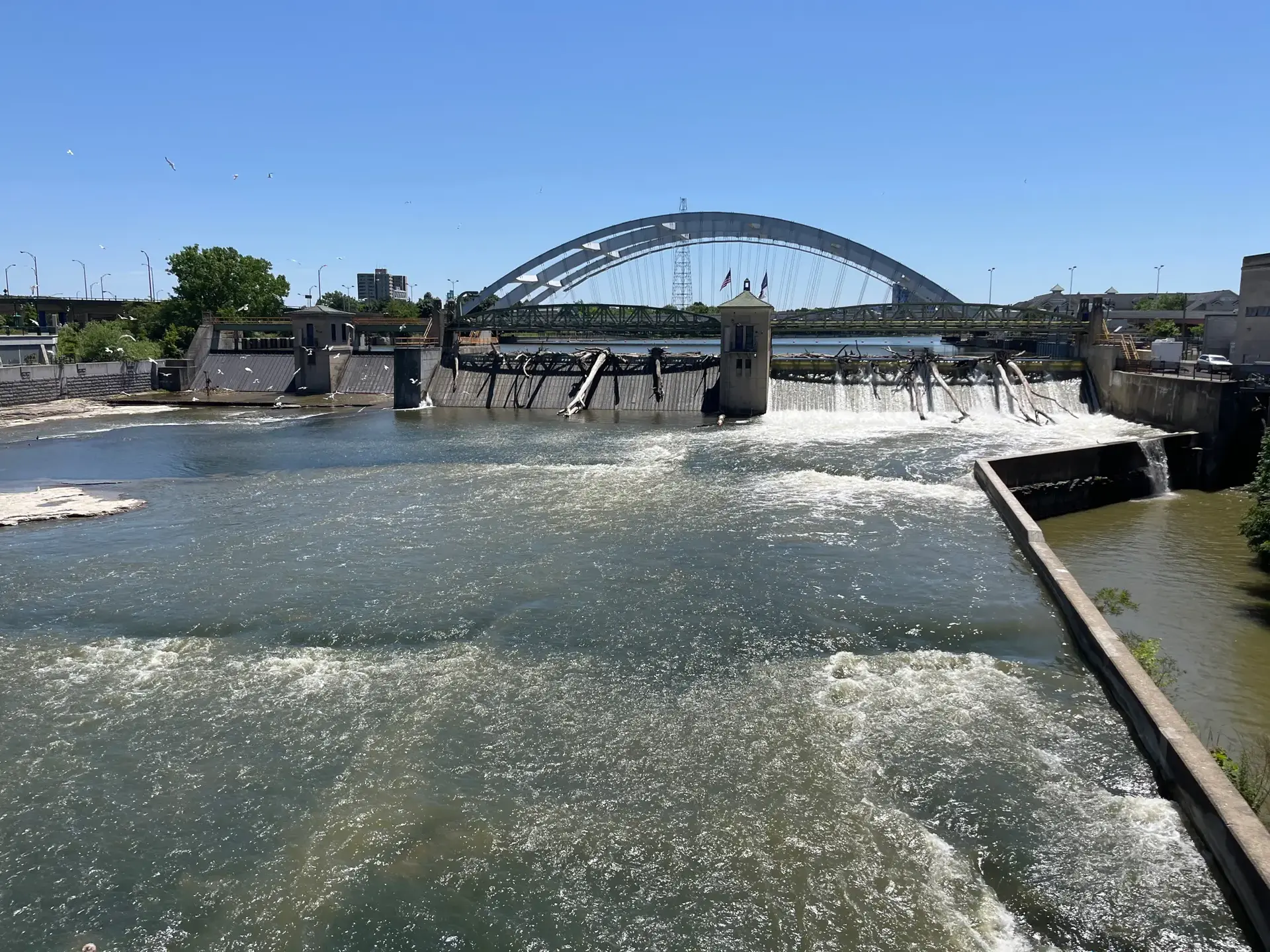 Bright sunny day over a dam with cascading water and an arched bridge.