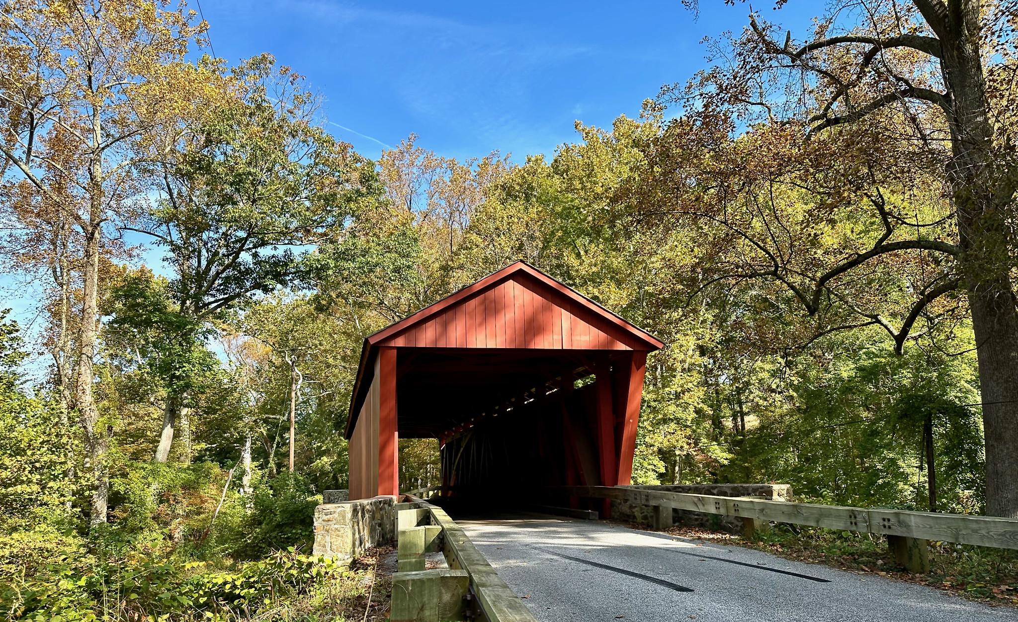 A red covered bridge sits among autumn trees on a sunny day.