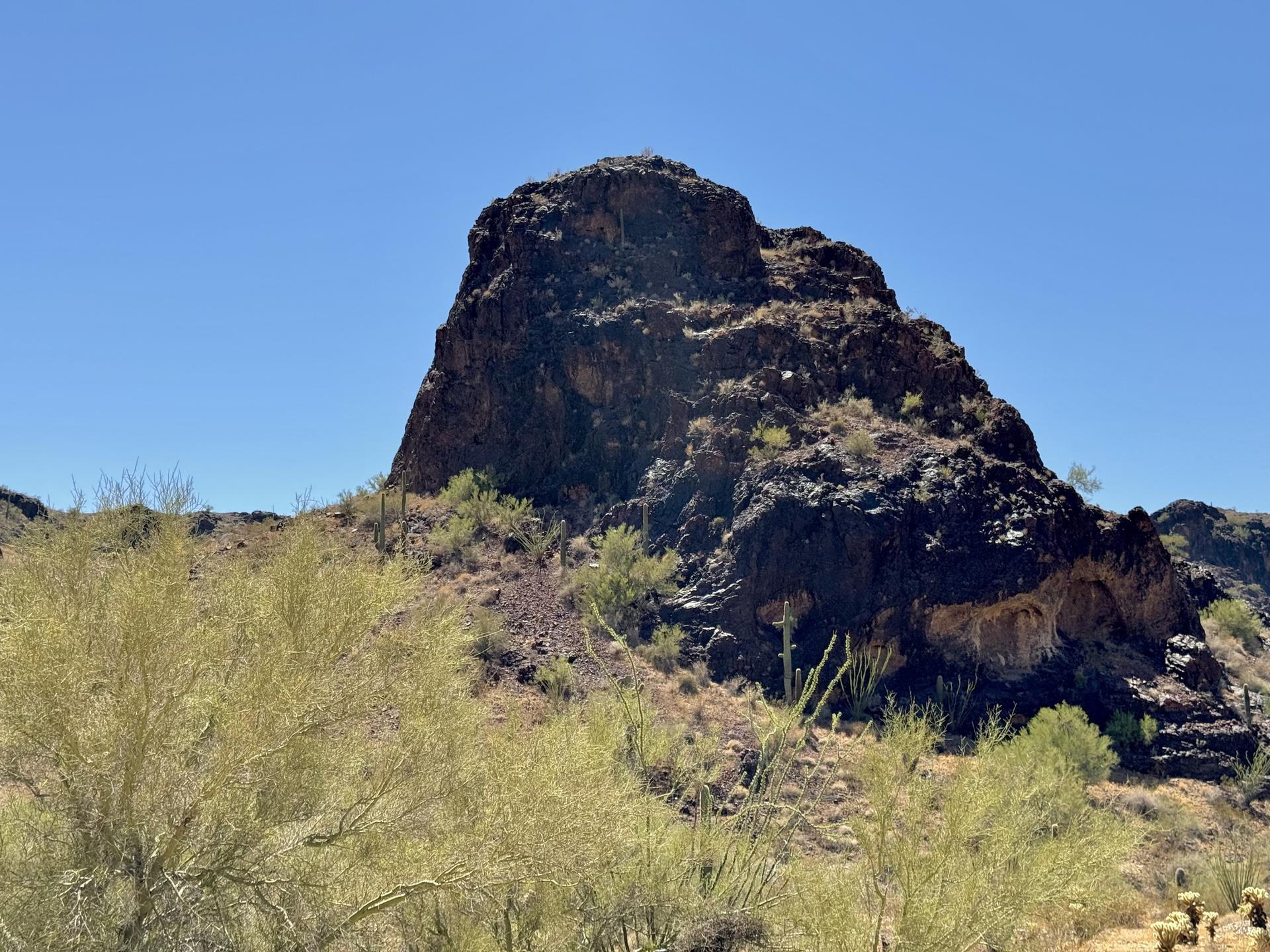 Large dark rock formation in a desert landscape under a clear blue sky.