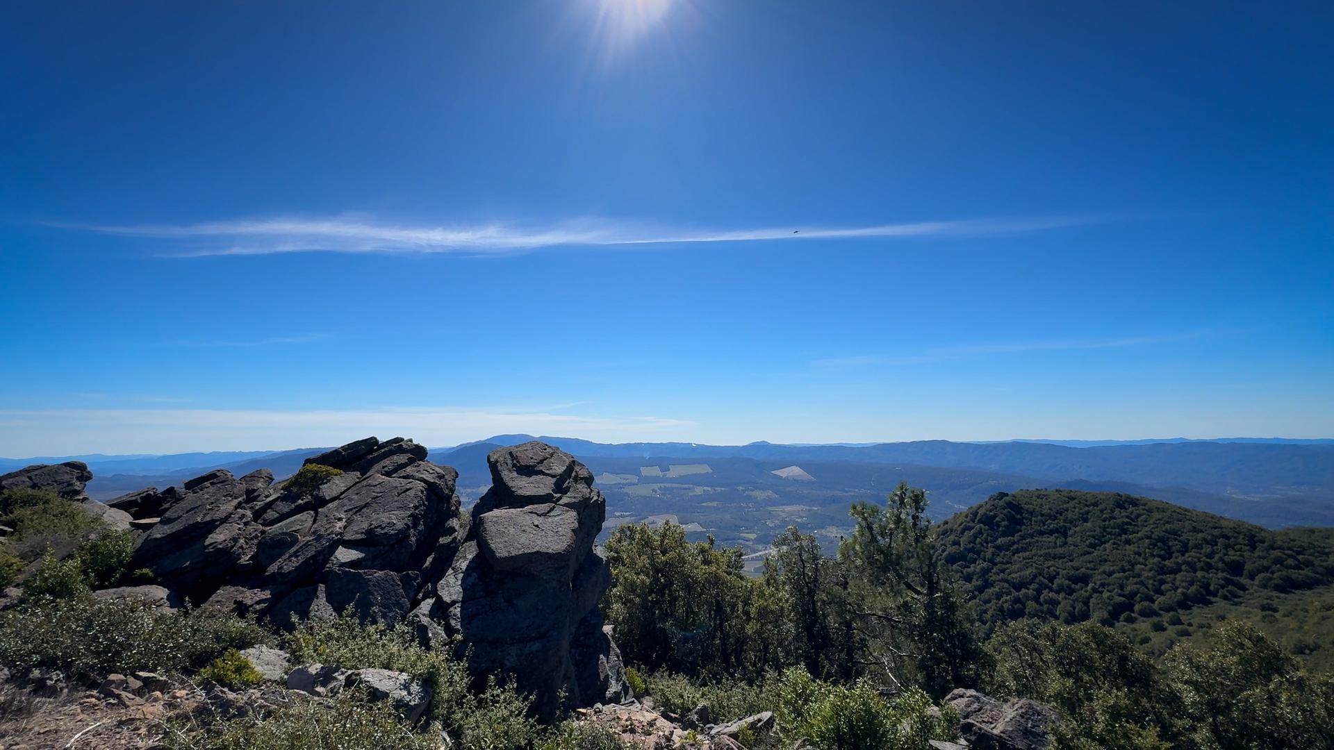 Sunny mountain peak with large rocks overlooking green hills and a vast landscape.