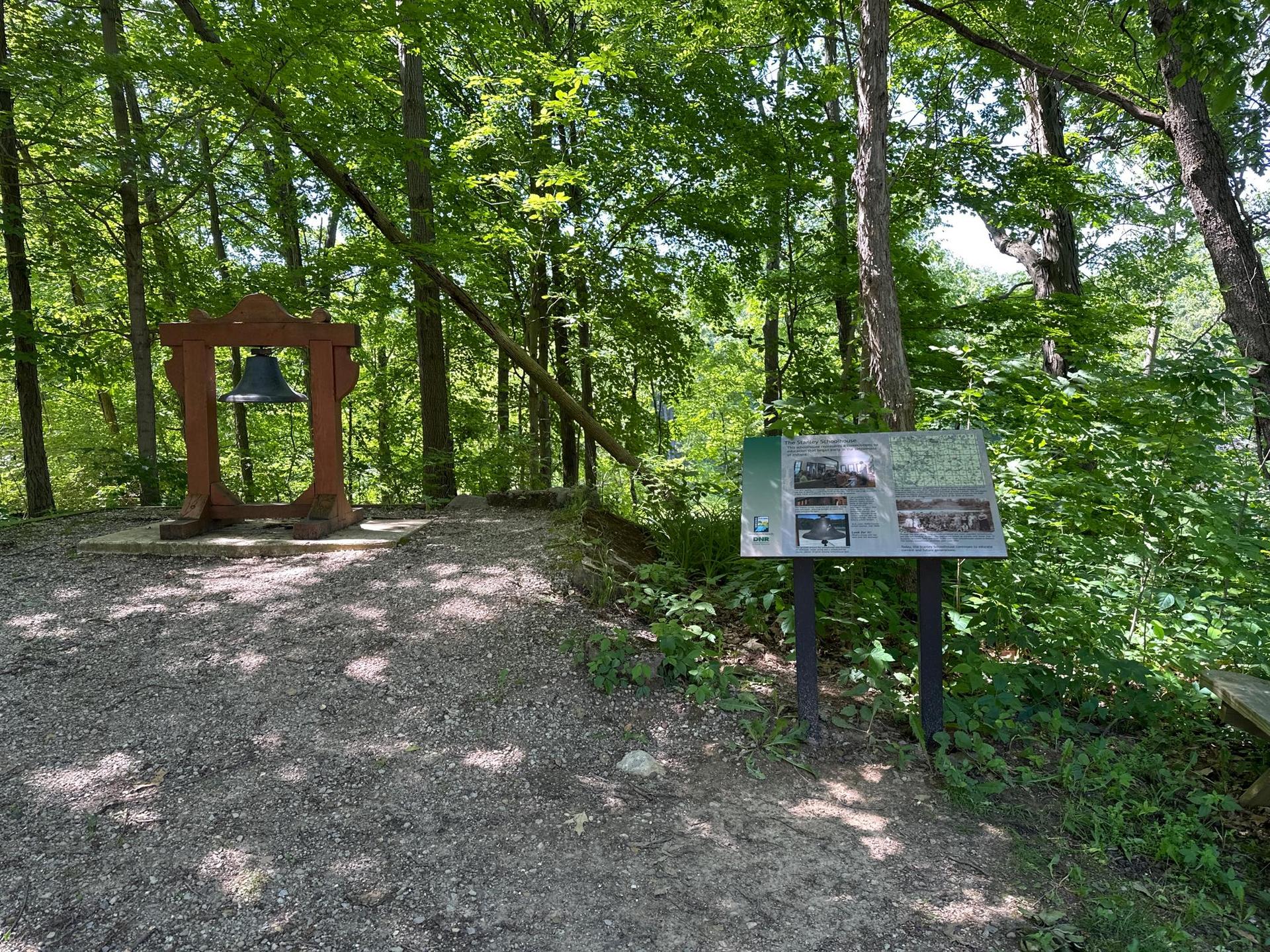 A large bell in a wooden frame and an informational sign in a sunlit forest.