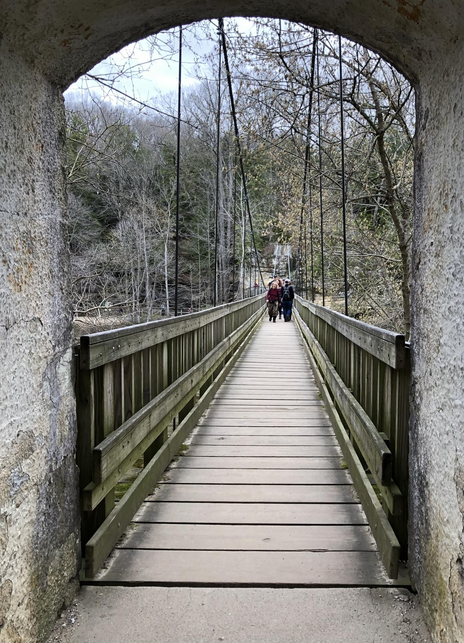 People walk on a wooden suspension bridge through a stone archway in a forest. - Turkey Run Trails 3, 10, 9, and 5 Loop