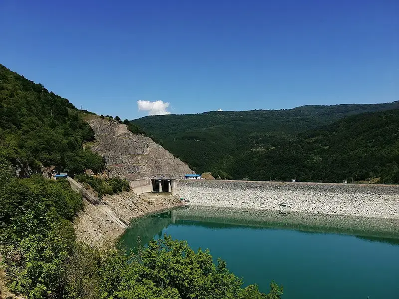 A large dam and turquoise reservoir surrounded by green mountains under a clear sky.