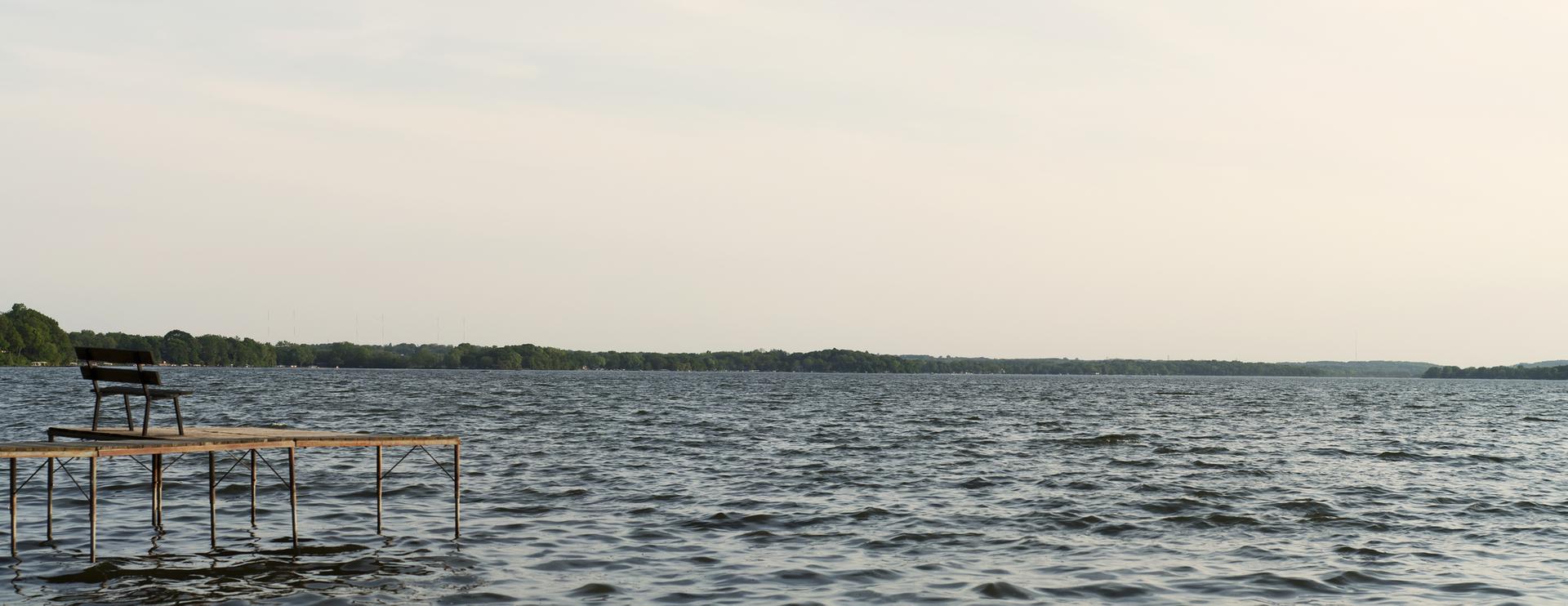 Bench on a wooden dock overlooking a wide lake with distant trees.