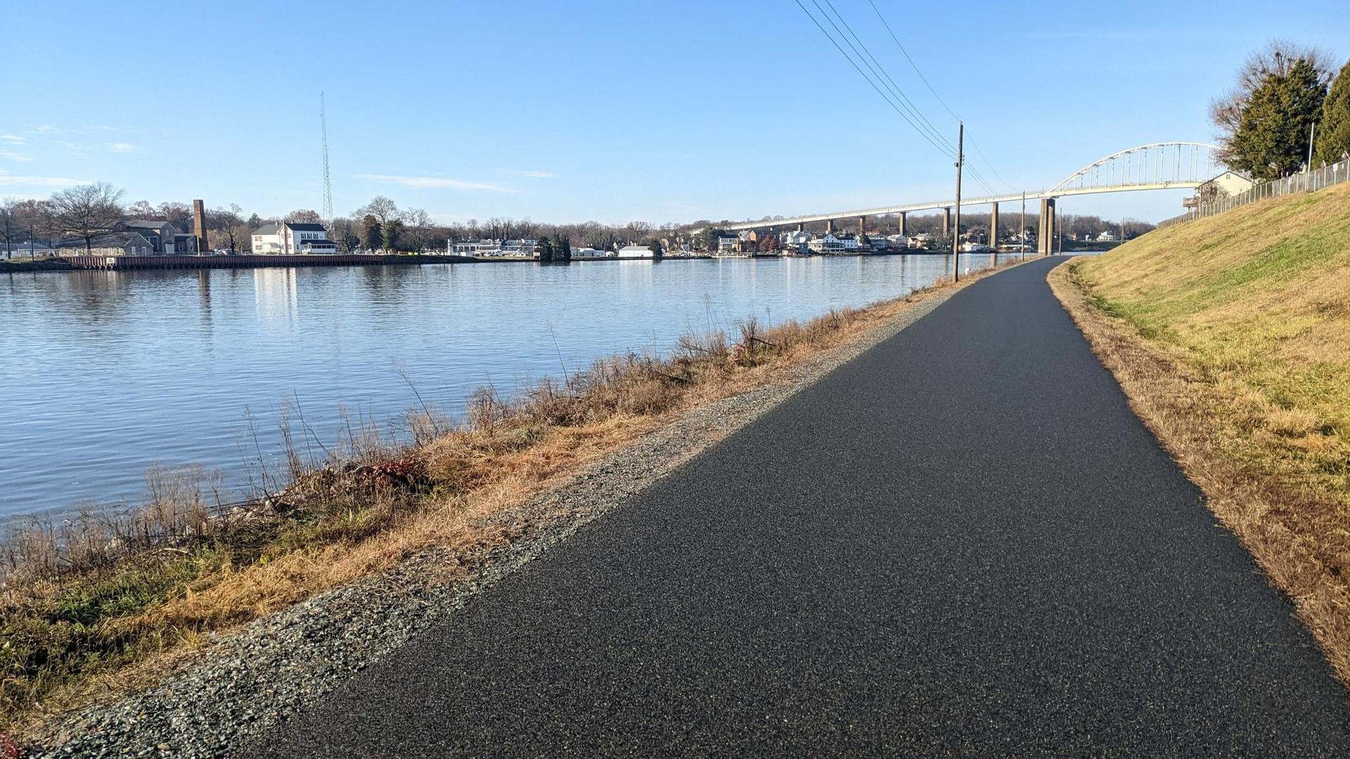 Paved path beside a calm river with a distant bridge on a clear day.