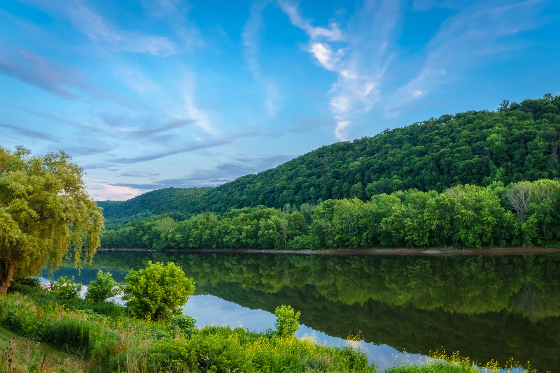 Calm river reflects blue sky and lush green hills under bright light.