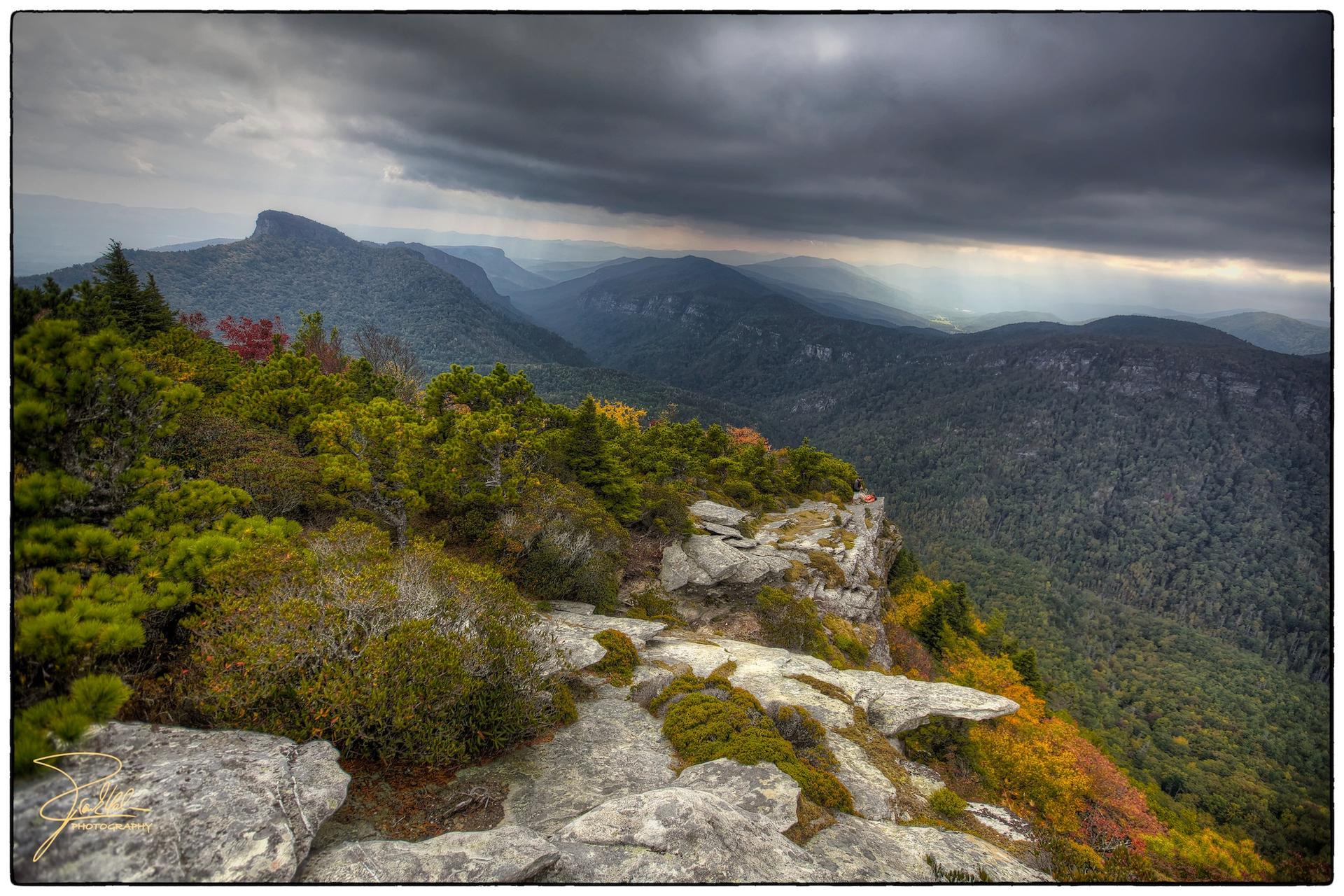 Rocky cliff overlooking dark, forested mountains under a dramatic stormy sky.