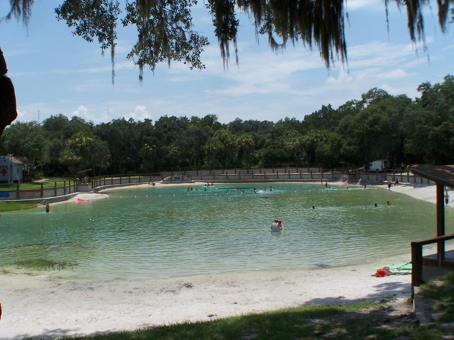 A sunny outdoor swimming area with people in clear water and sandy beach.