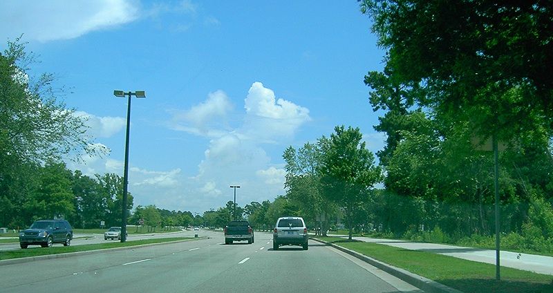 Cars drive on a multi-lane road lined with trees under a bright blue sky. - Grissom Parkway Trail