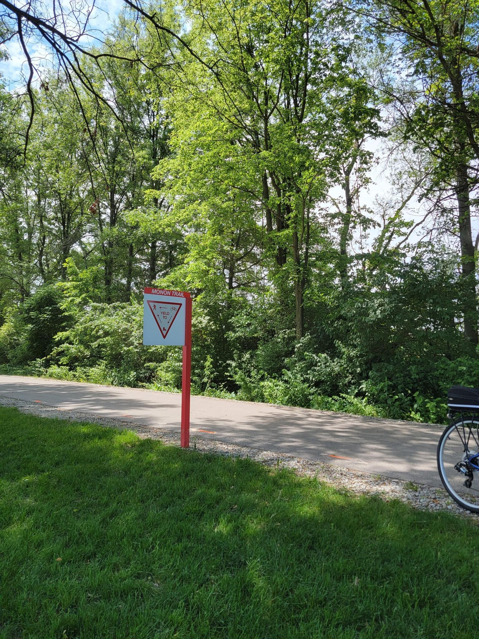 A red and white sign beside a paved path, with green trees and a bicycle wheel.