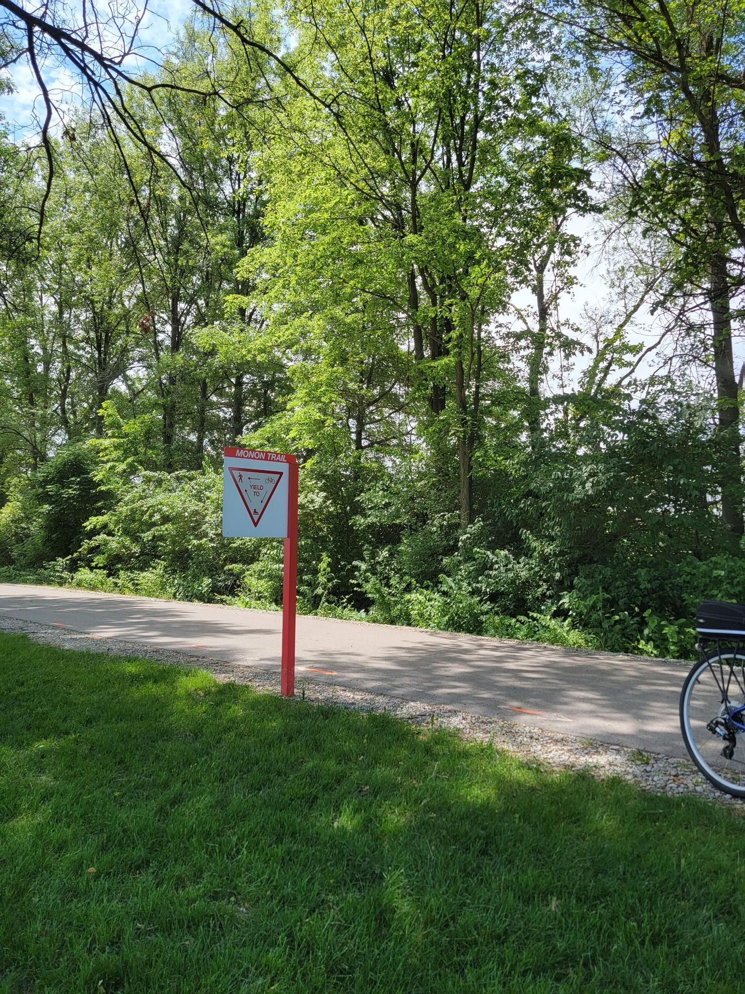 A red and white sign beside a paved path, with green trees and a bicycle wheel. - Monon Trail Loop from Carmel