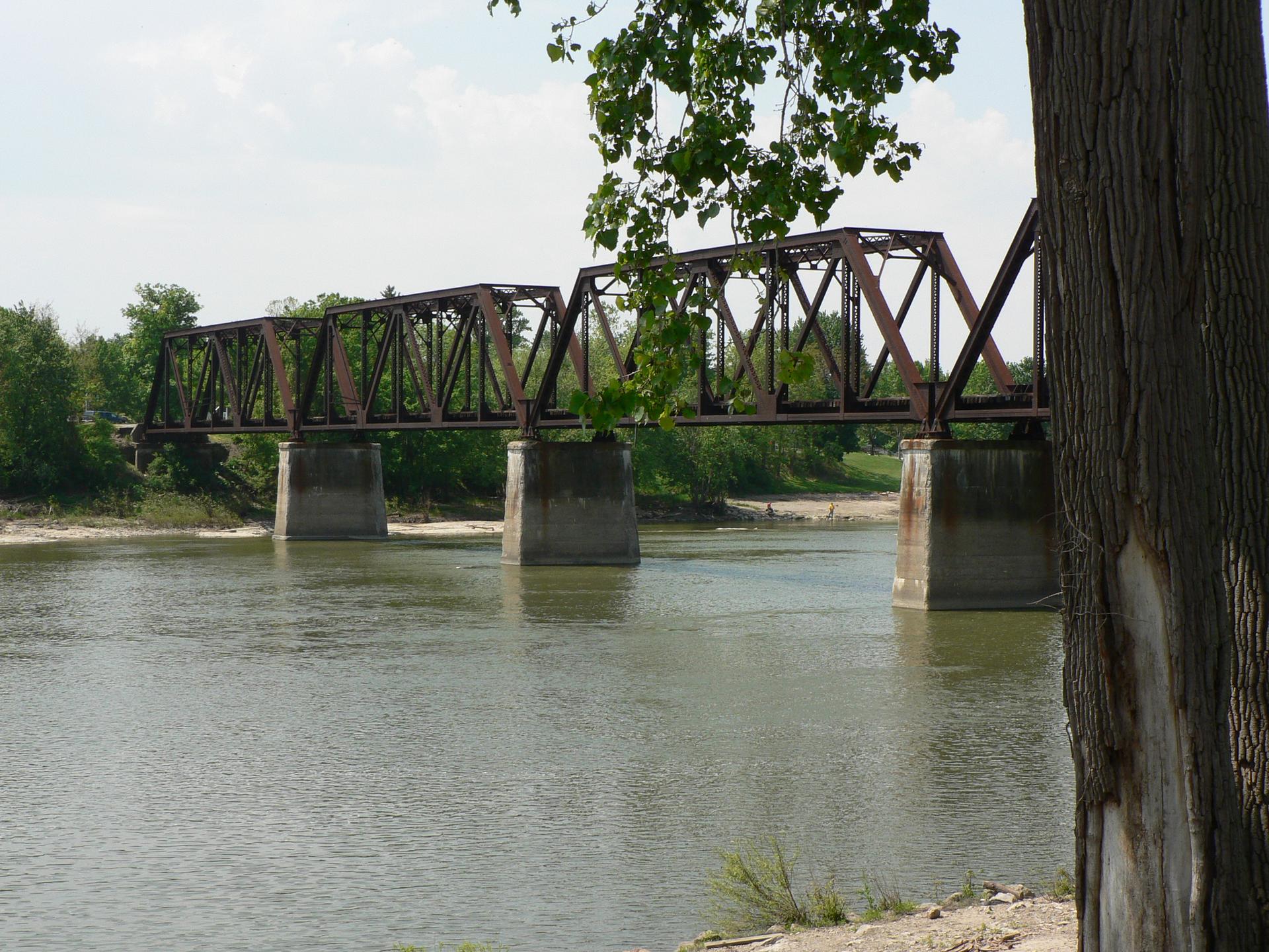 A long metal truss bridge spans a wide river on a bright day.