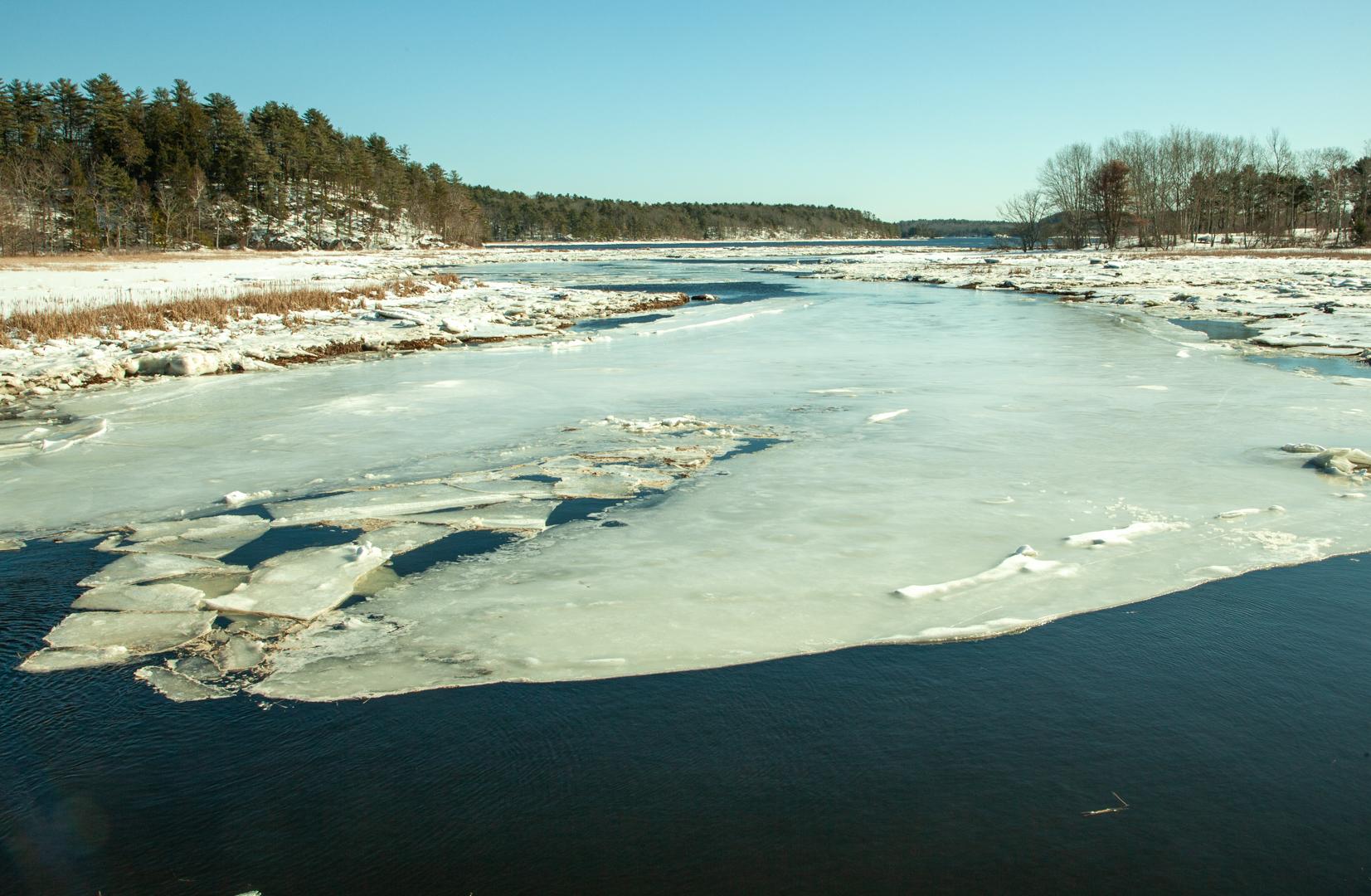 Wide river partially frozen with ice, snow-covered banks, and distant trees.