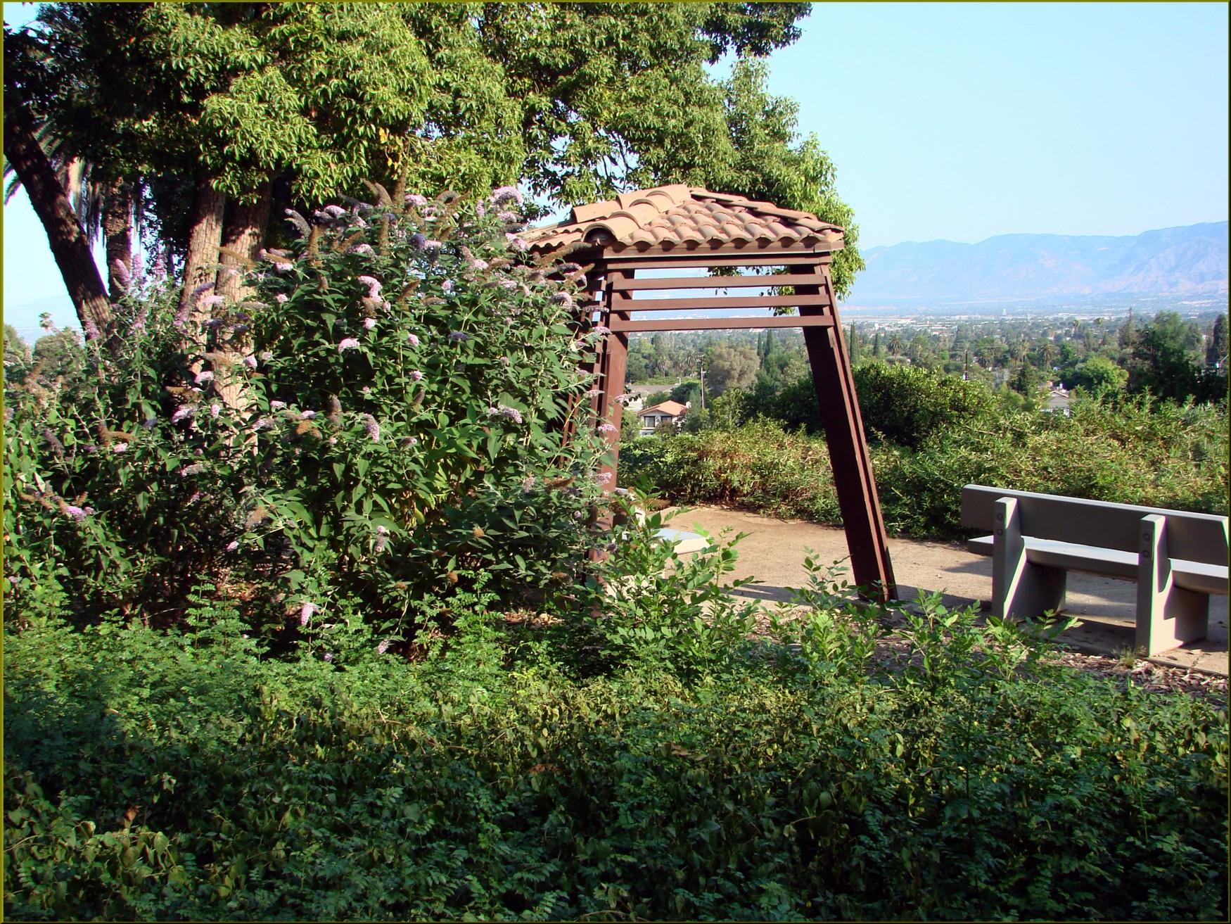 Sunny overlook with a tiled shelter, benches, and green foliage, distant mountains.