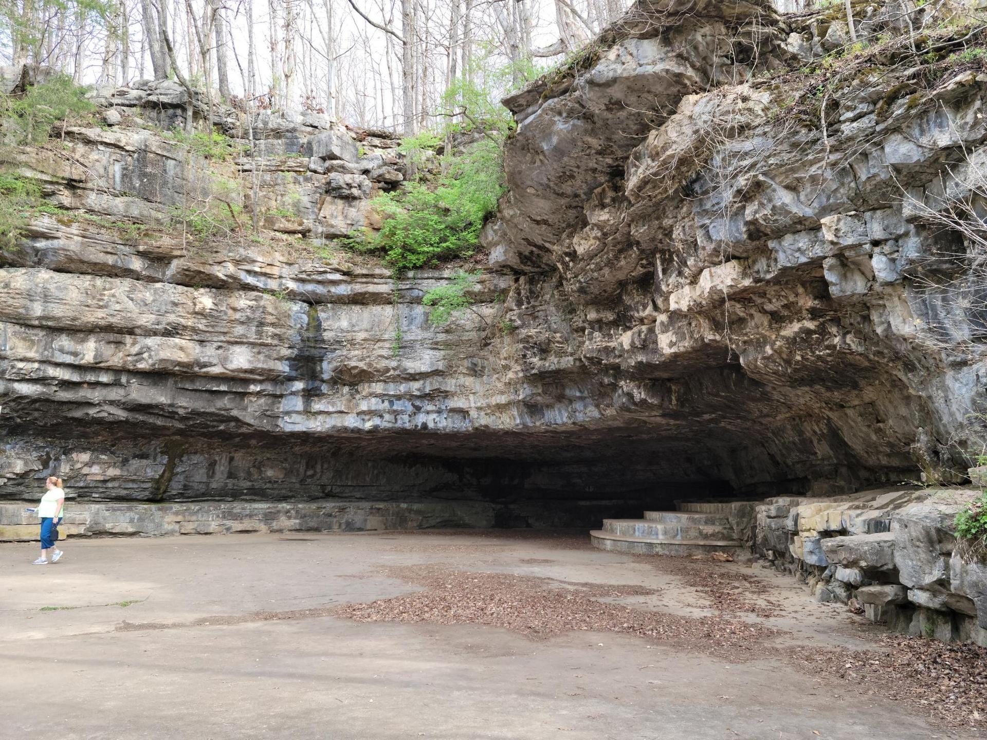 A person stands before a large, dark cave entrance in a rocky cliff.