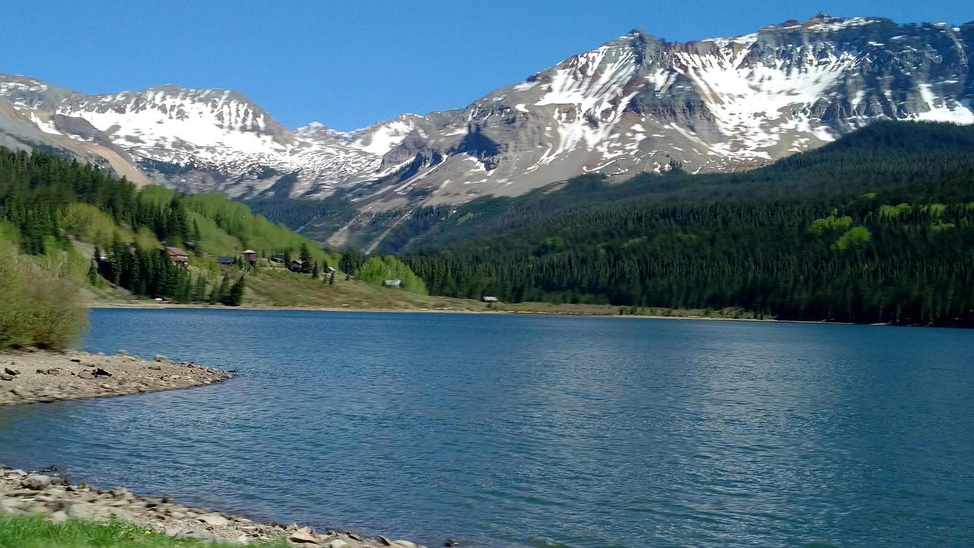 A clear lake reflects snow-capped mountains and green forests under a blue sky.