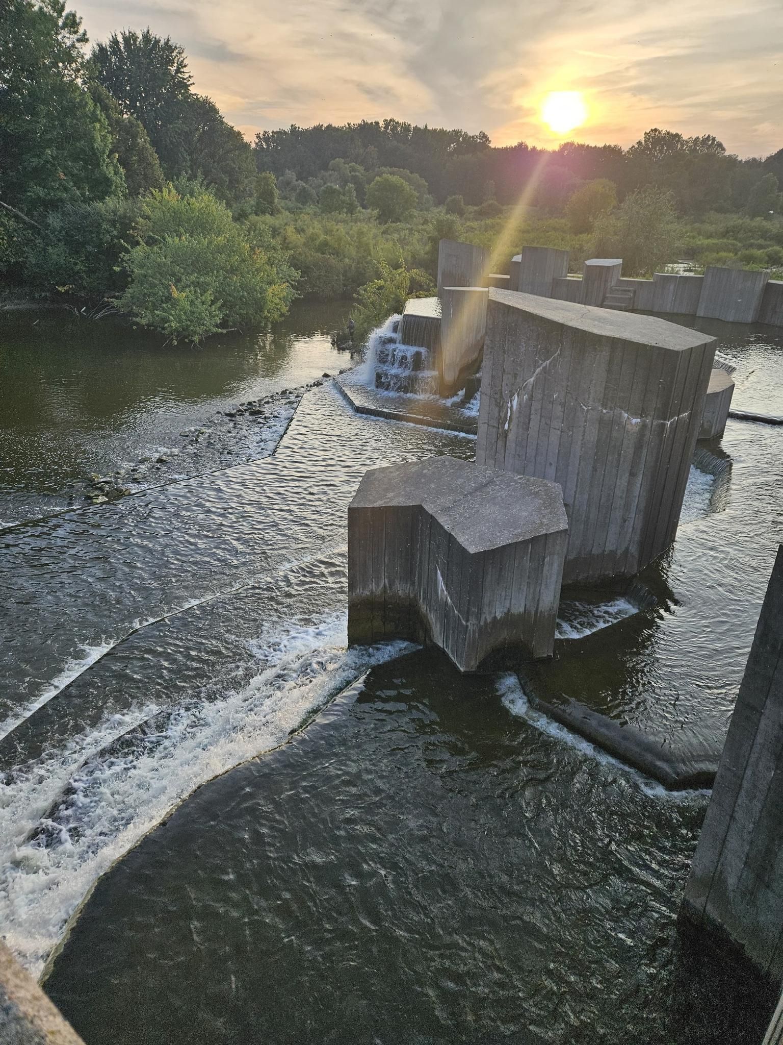 Sun flares over a river flowing through a concrete dam structure with trees. - Flint River Trail