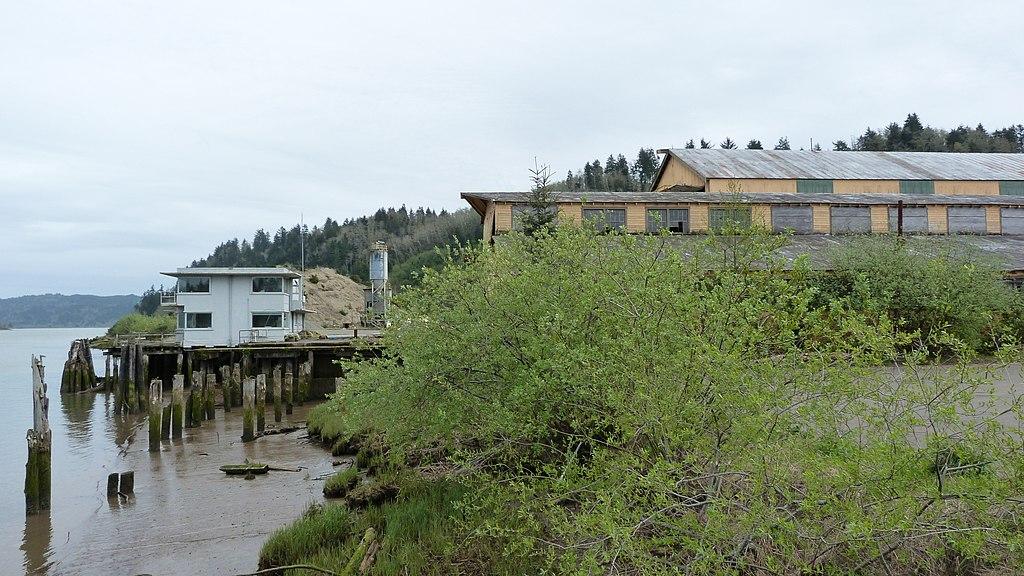 Old buildings on stilts over murky water, with a forested hill.