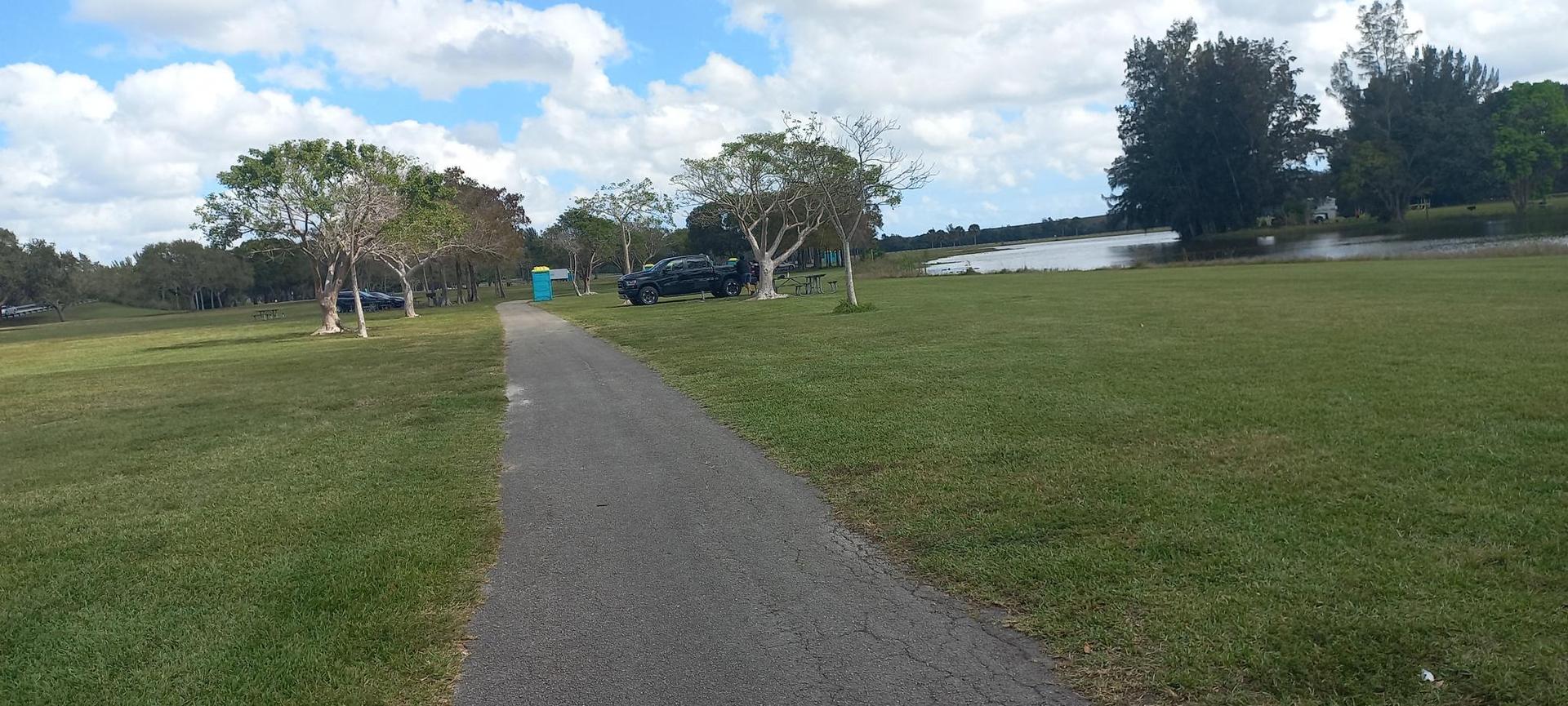 Paved path through a green field with scattered trees and a distant lake.