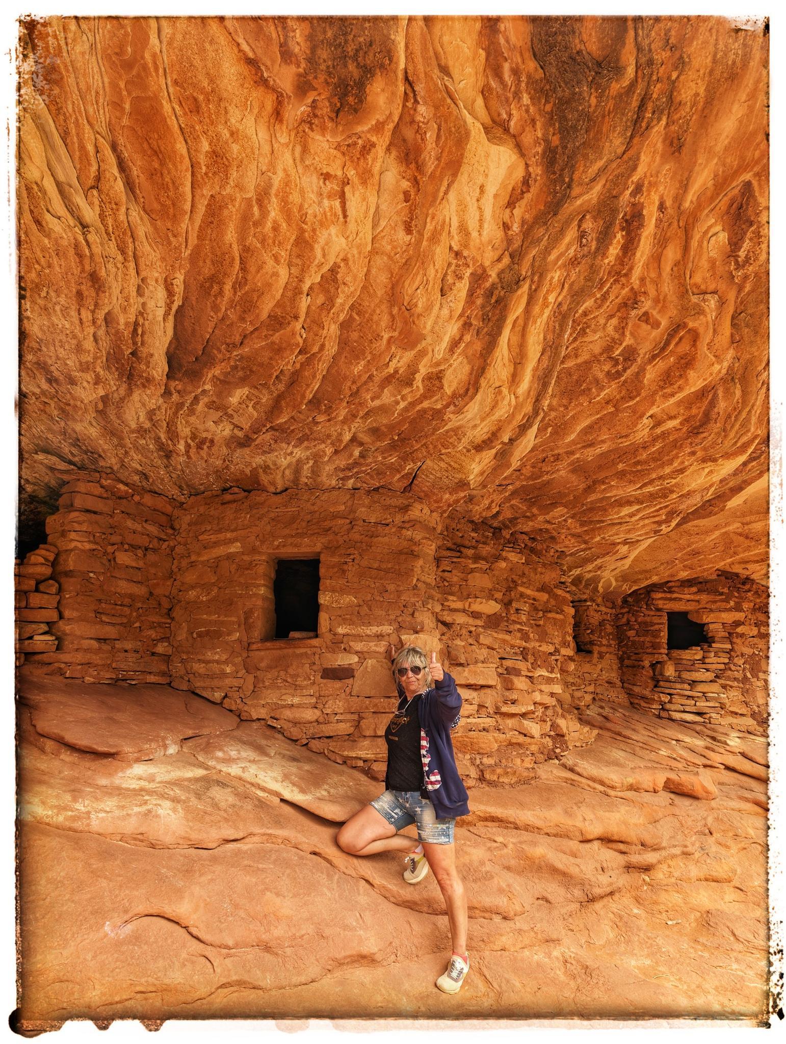 Woman poses before ancient stone dwellings built into a massive orange rock overhang.