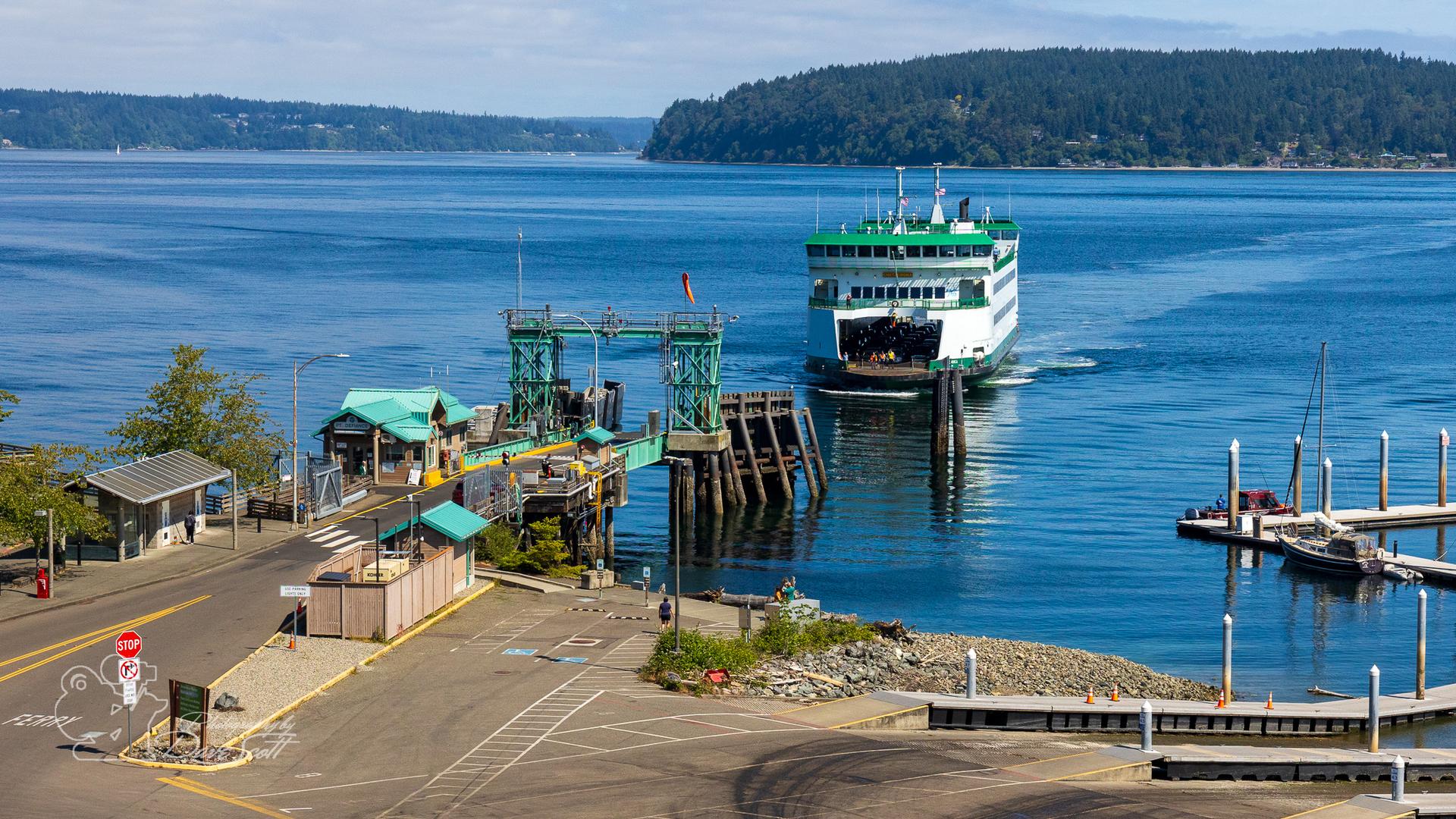 Bright ferry boat approaches a dock on blue water with forested land.