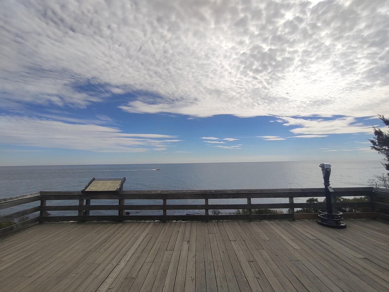 Wooden observation deck with telescope overlooks a vast, cloudy ocean horizon.