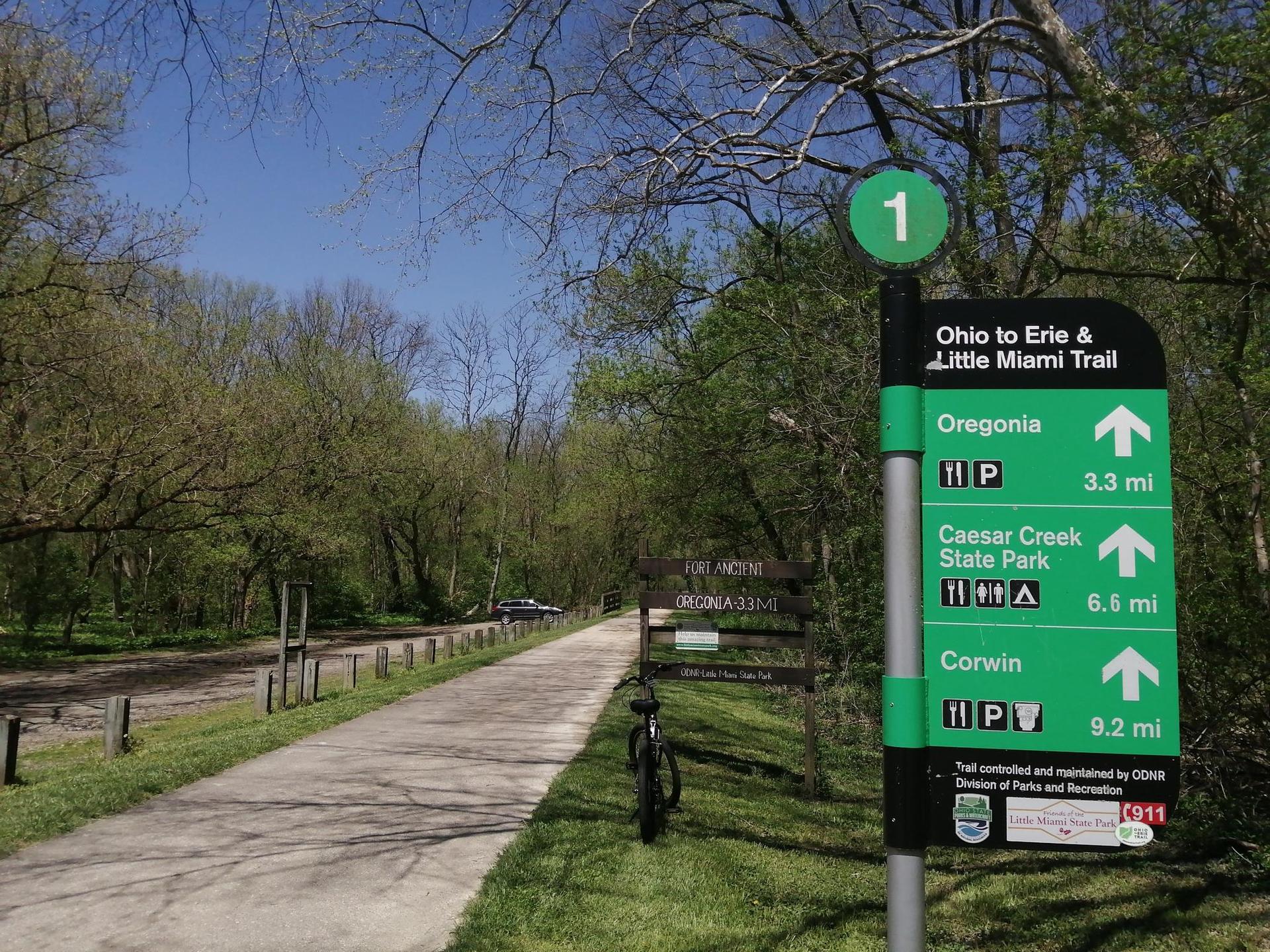 Bright daylight illuminates a green trail sign next to a paved path through a wooded area.