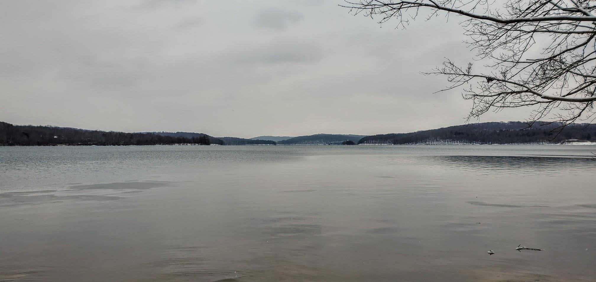 Wide, calm lake surrounded by snow-dusted hills under an overcast sky.