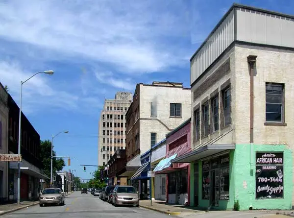A sunny urban street lined with various storefronts and parked cars.