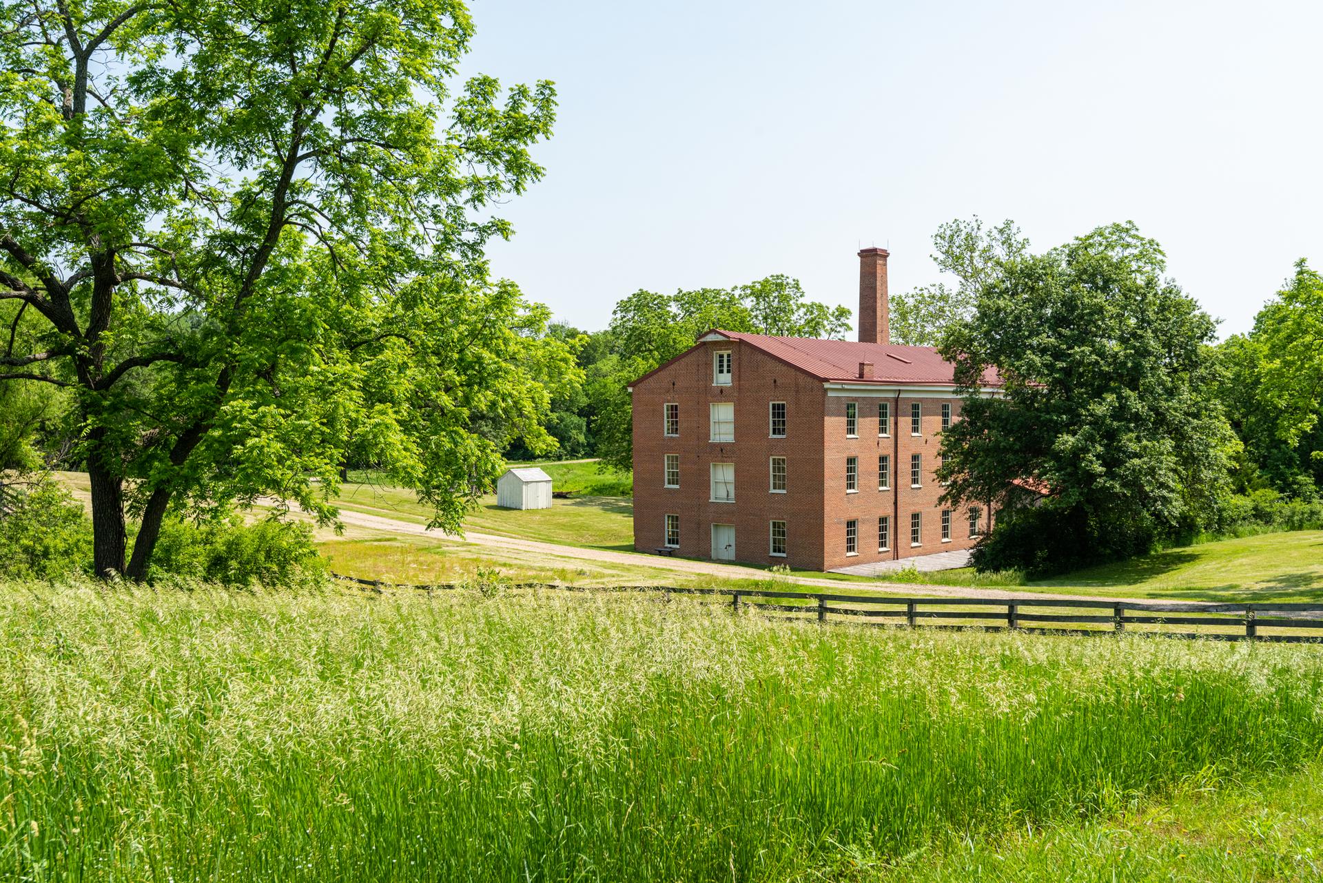 A large red brick building surrounded by green fields and trees on a sunny day.