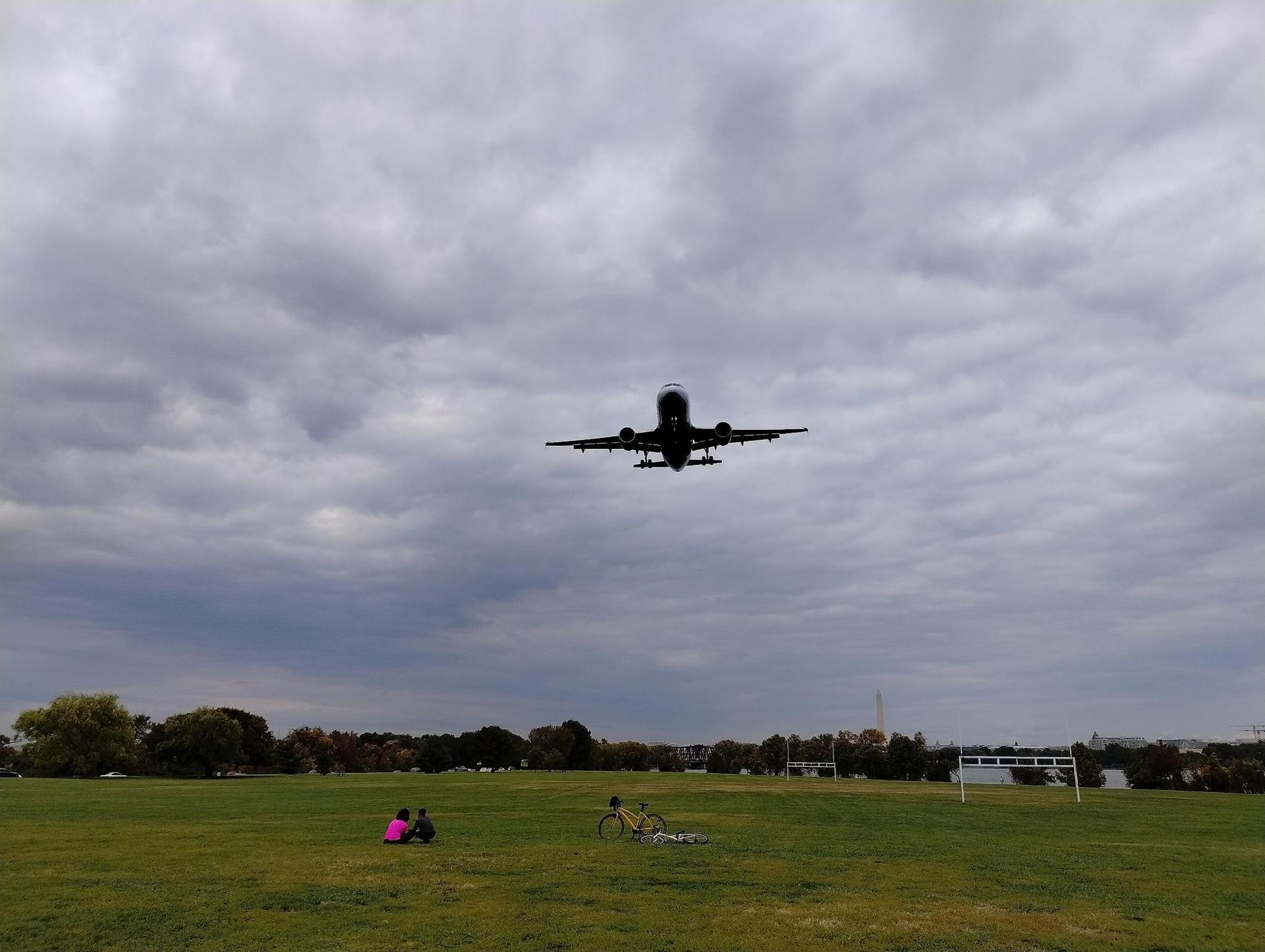 Large airplane descending over a green field with two people sitting.