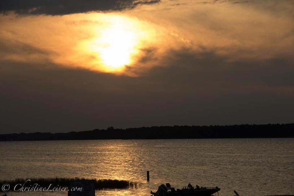 Golden sunset over dark water with a silhouetted boat and distant trees.