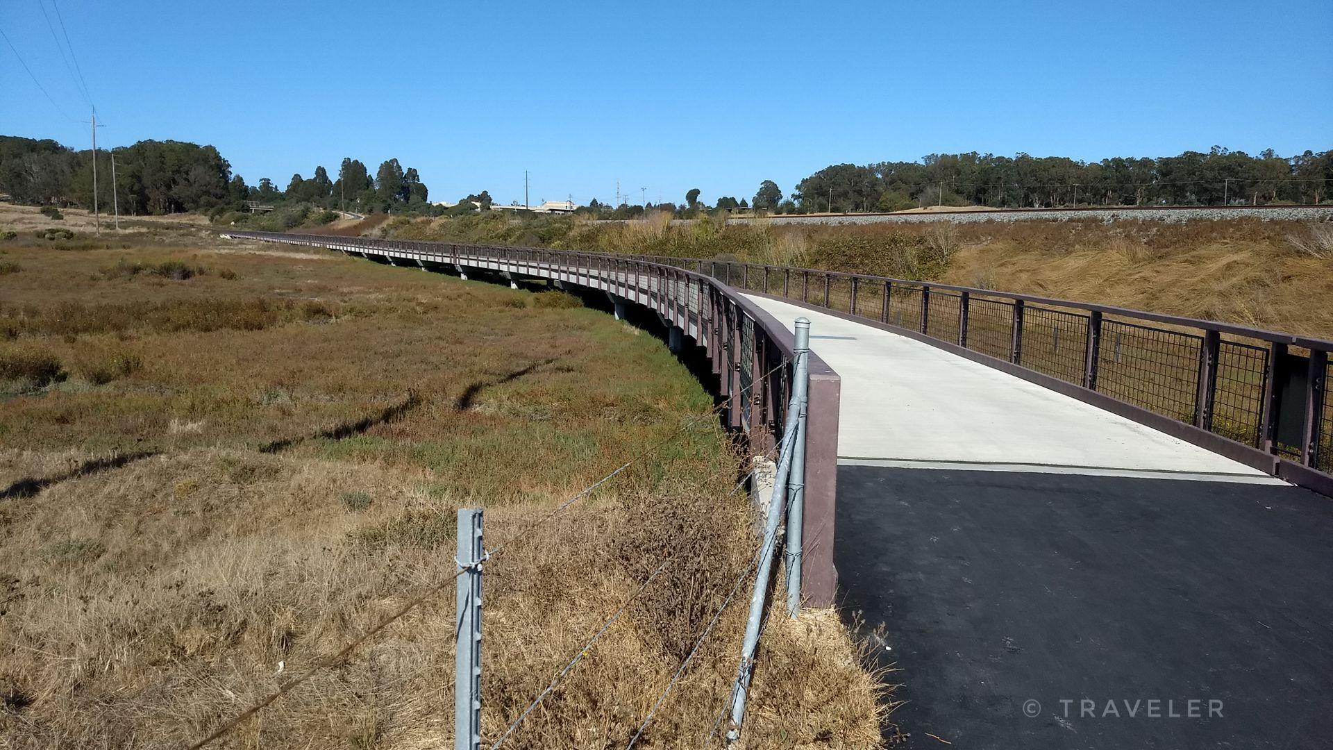 A long, curving boardwalk path extends through dry marshland on a sunny day.