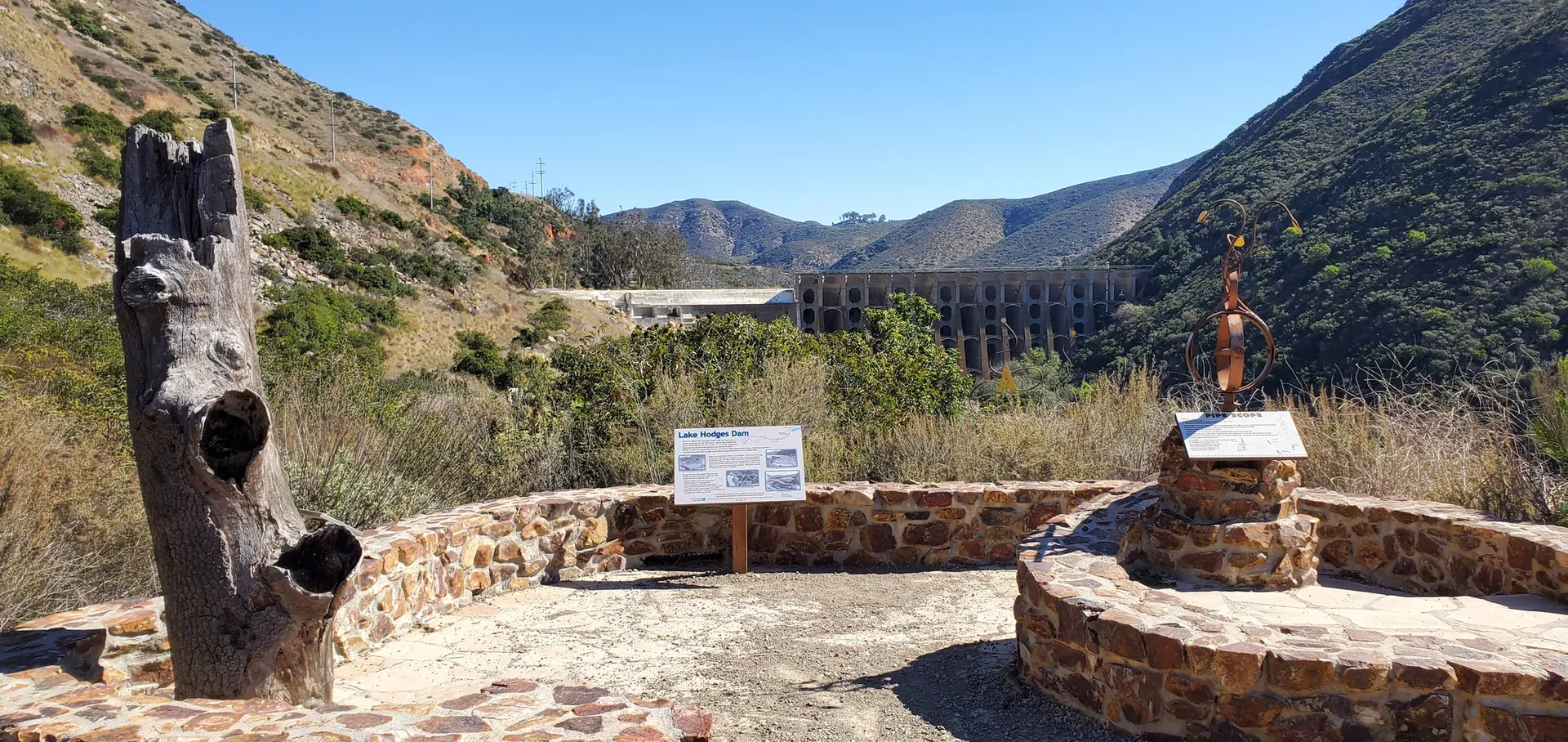 Sunny view of a dam from a stone viewing area with signs and sculpture.