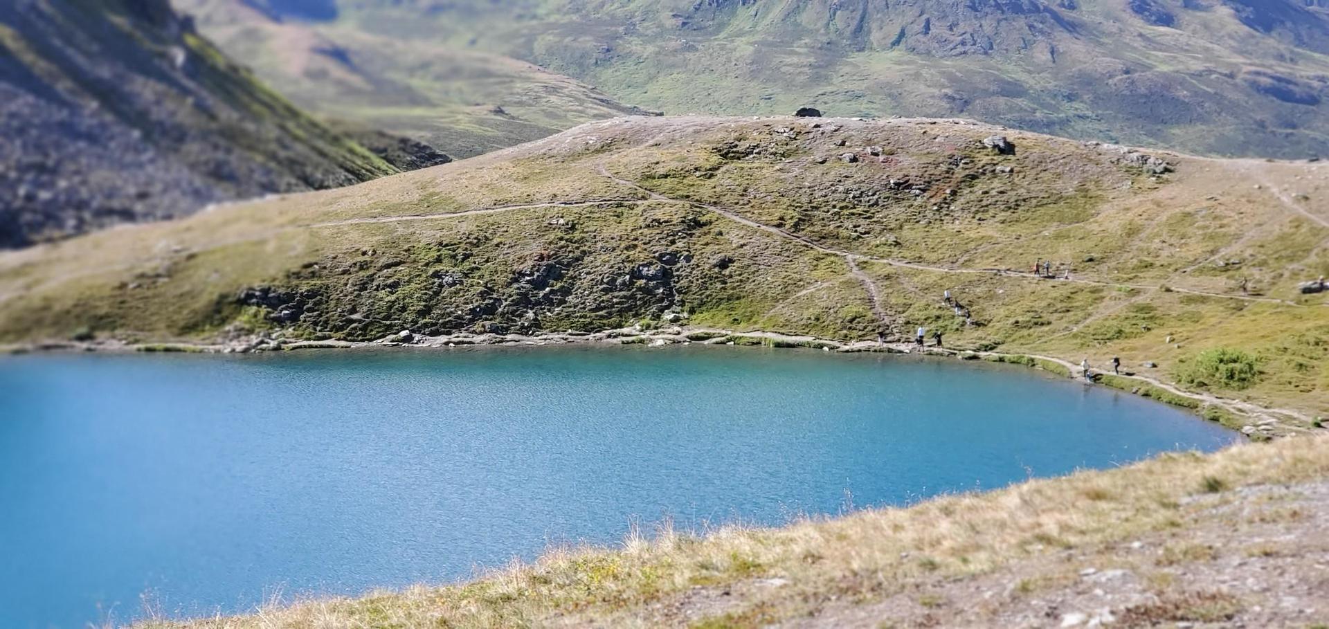 Bright blue lake nestled among grassy hills with distant mountains.