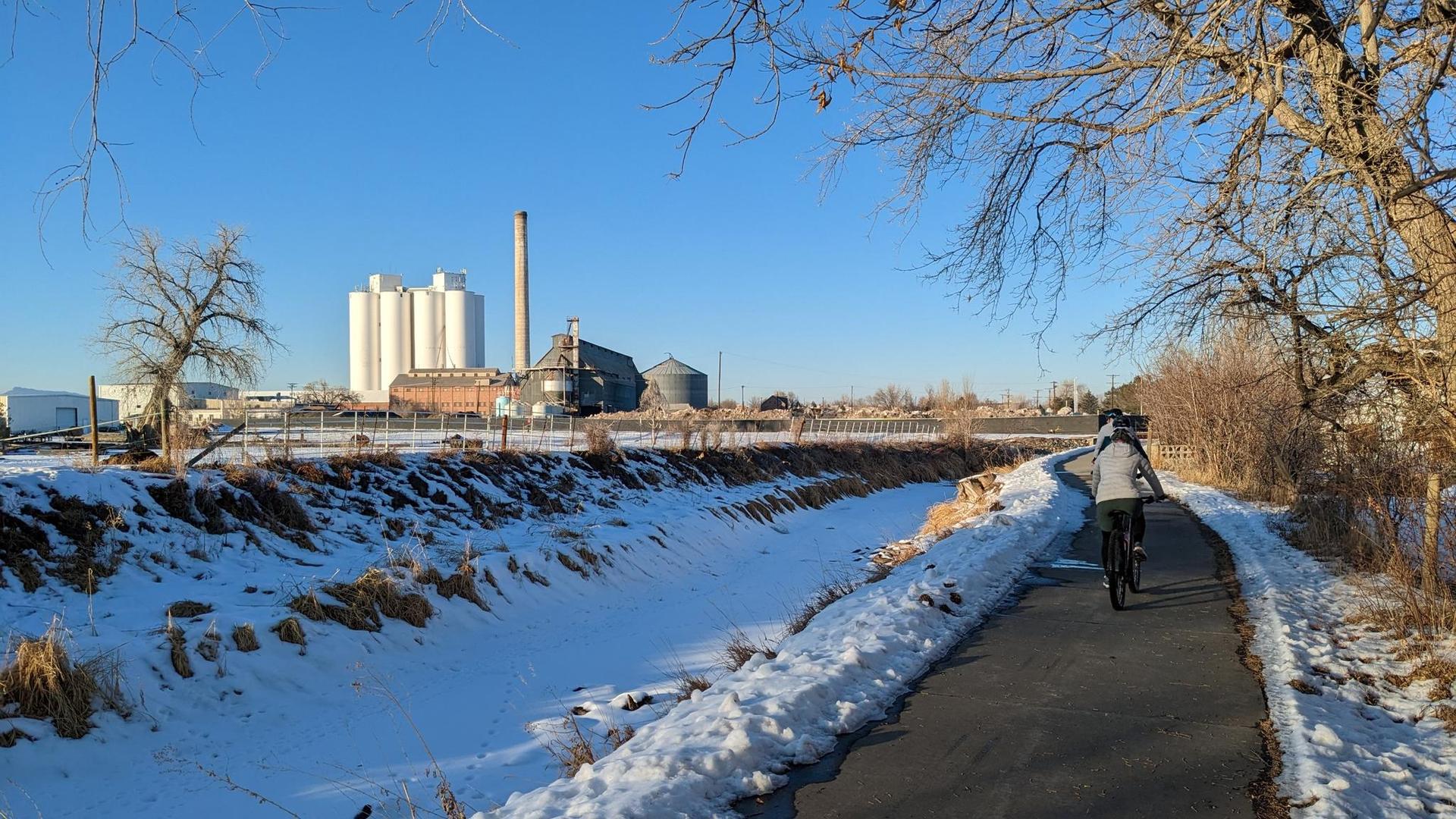 Cyclist on a paved path beside a snowy ditch, industrial buildings under blue sky.