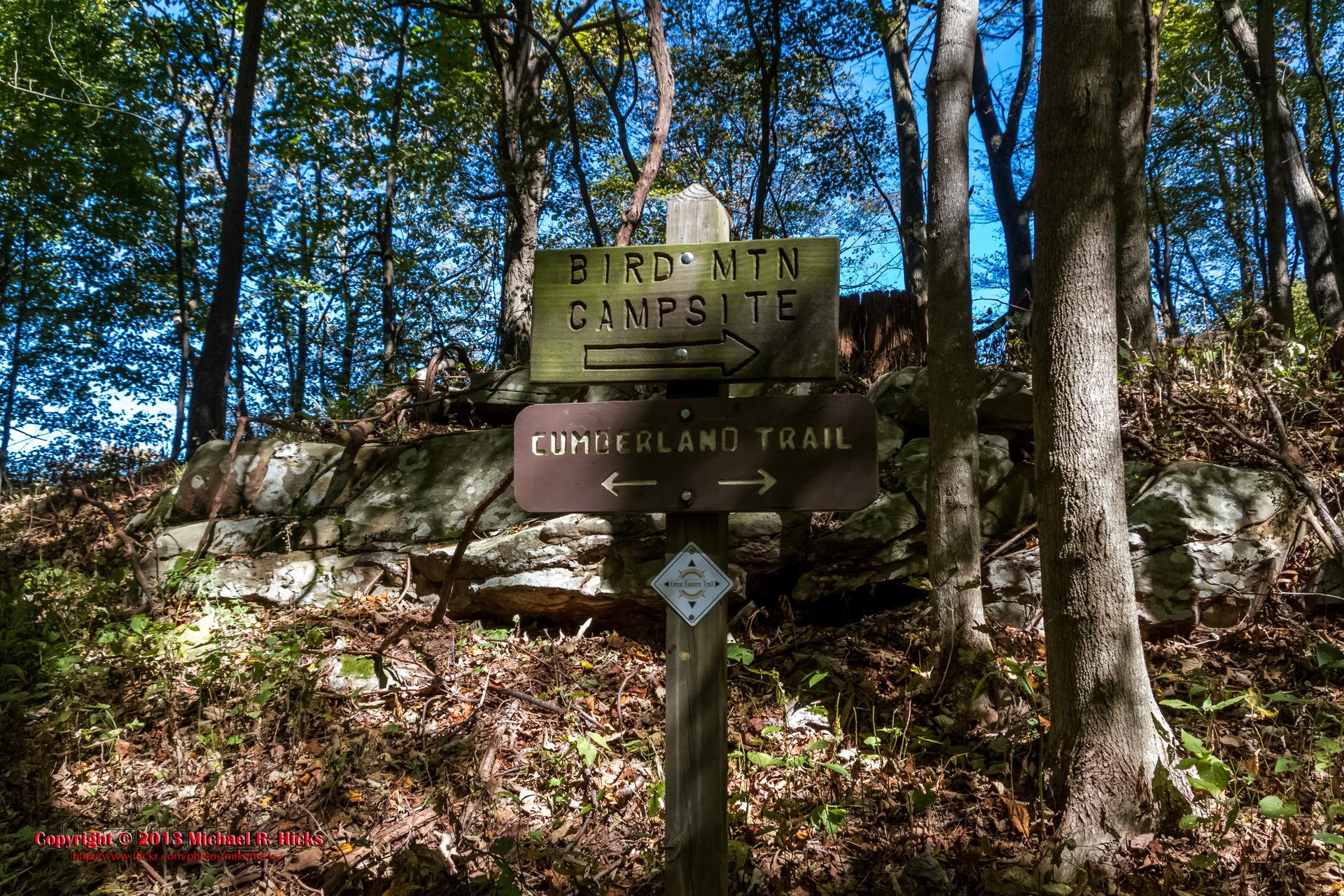 Two wooden trail signs stand in a forest with a rocky background.