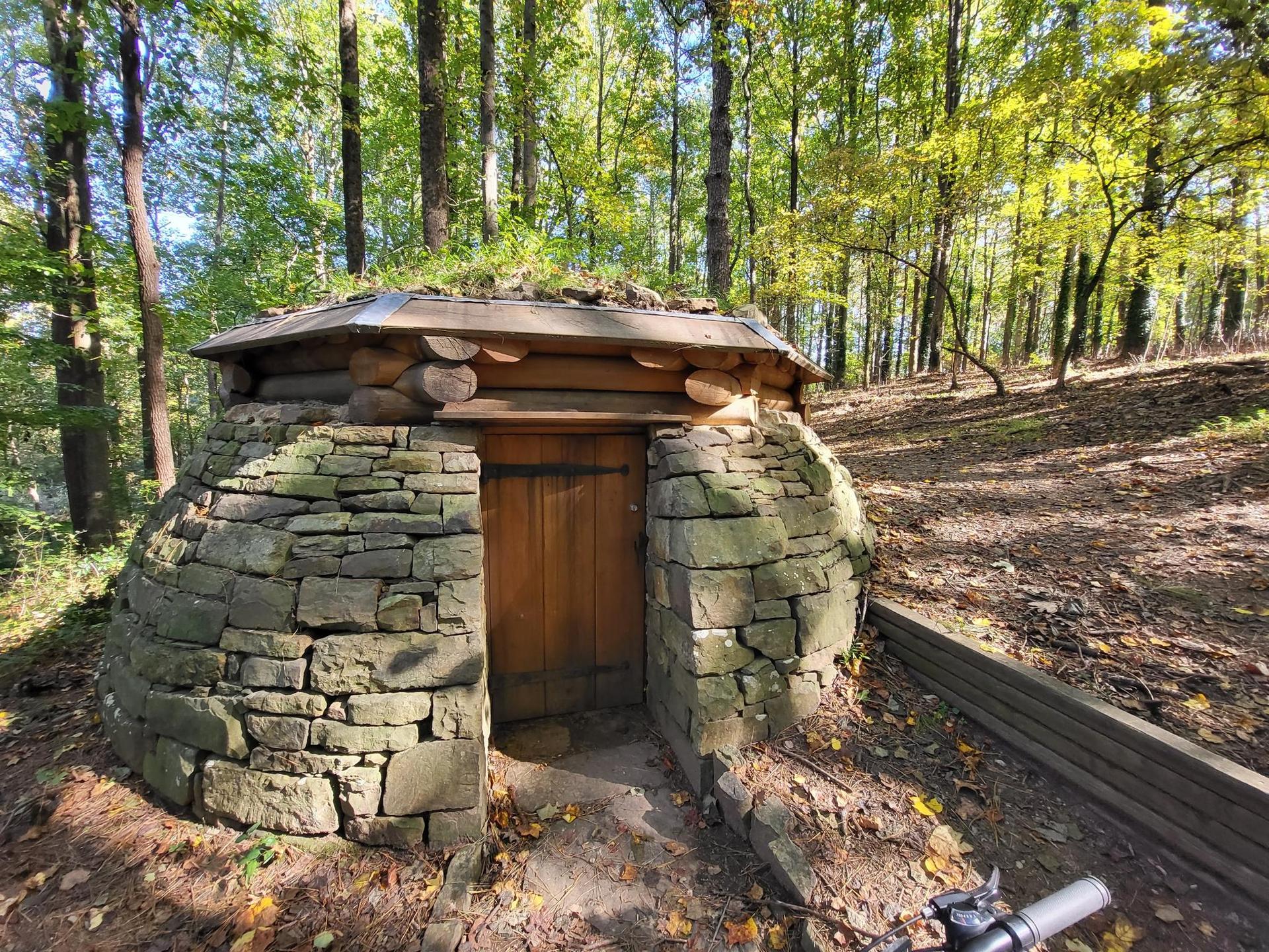 Stone and wood structure with moss roof in a sunny forest, bike in foreground.