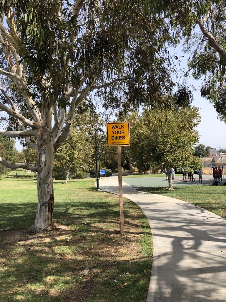 A "Walk Your Bikes" sign next to a paved park path with trees.