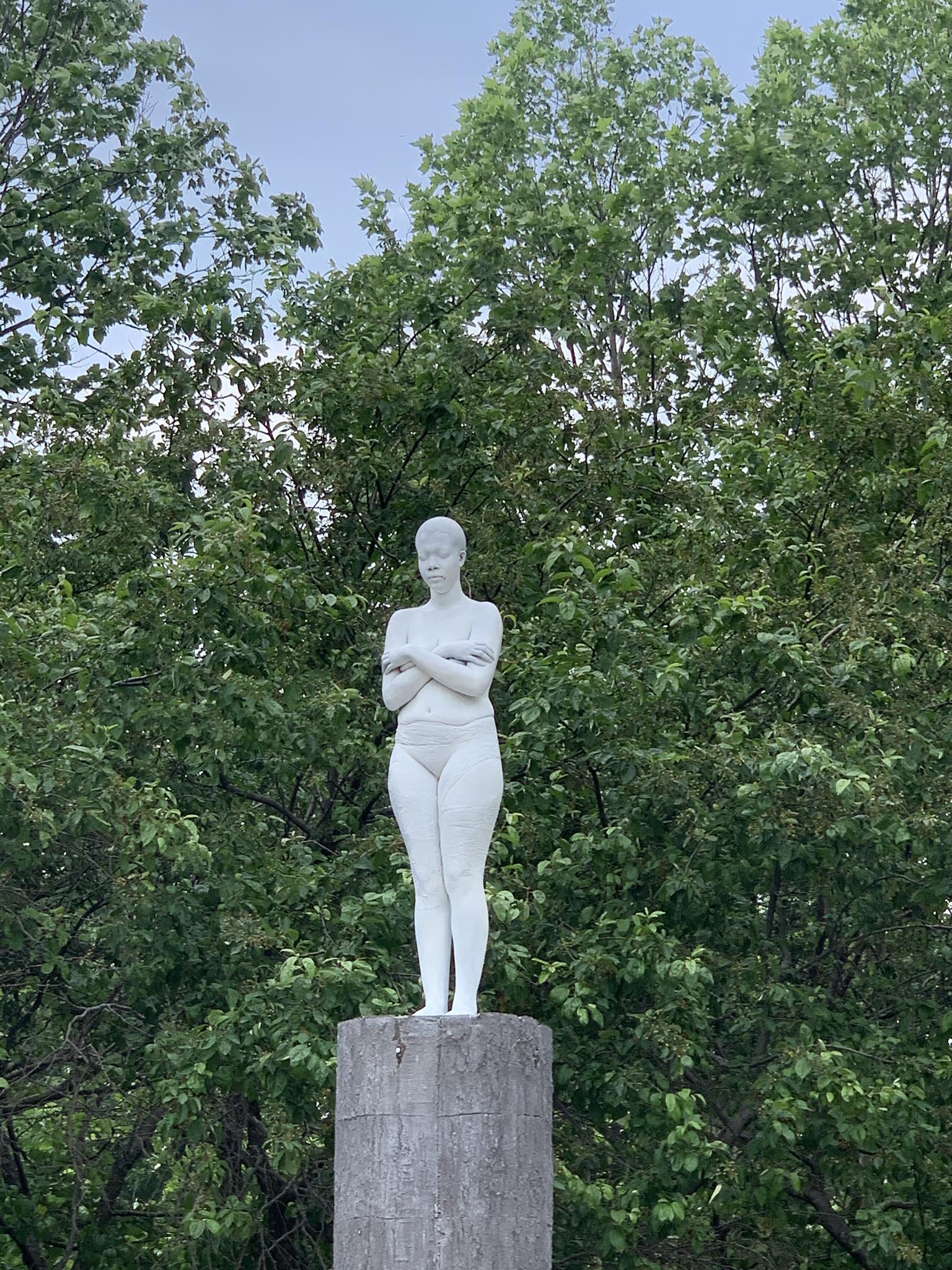 A white statue of a woman on a pedestal against green trees.