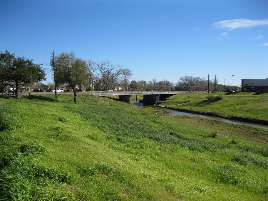 Sunny view of a bridge over a narrow stream with green grassy banks.