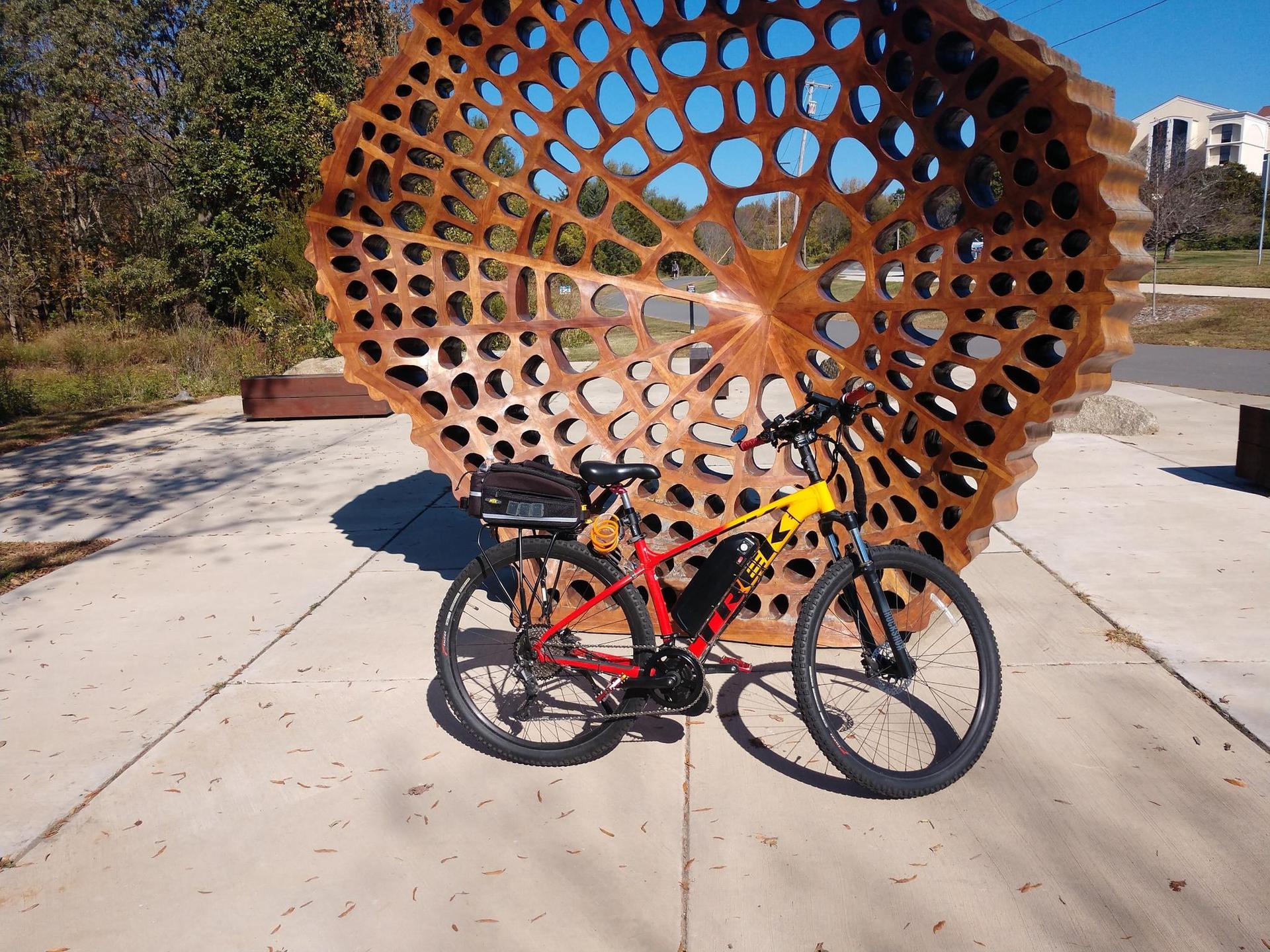 Electric bicycle parked next to a large, abstract, perforated sculpture on pavement.